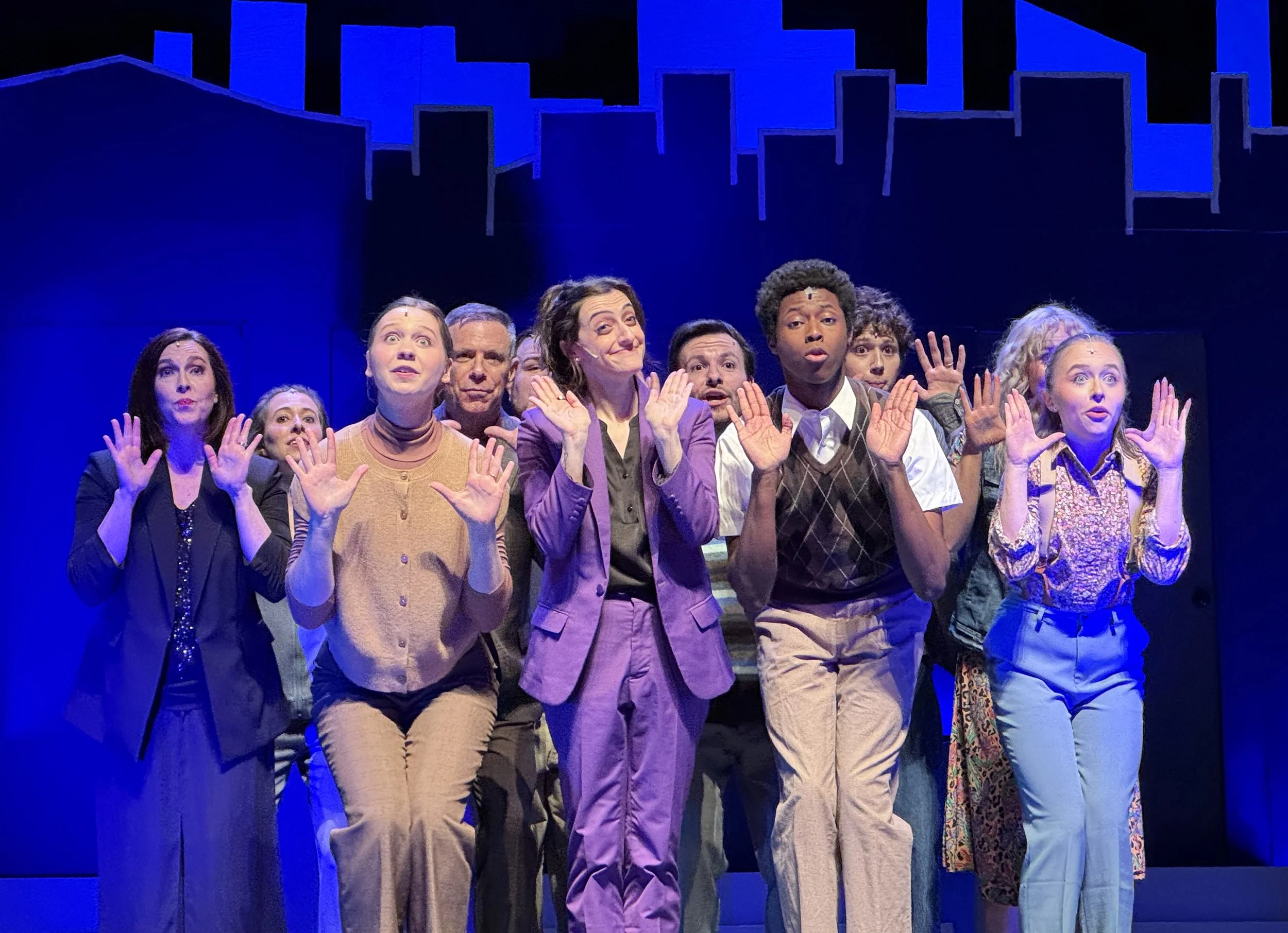 Group of nine actors on stage, dressed in various casual costumes, standing in front of a dark blue background with geometric blue props, performing a theatrical scene with hand gestures.