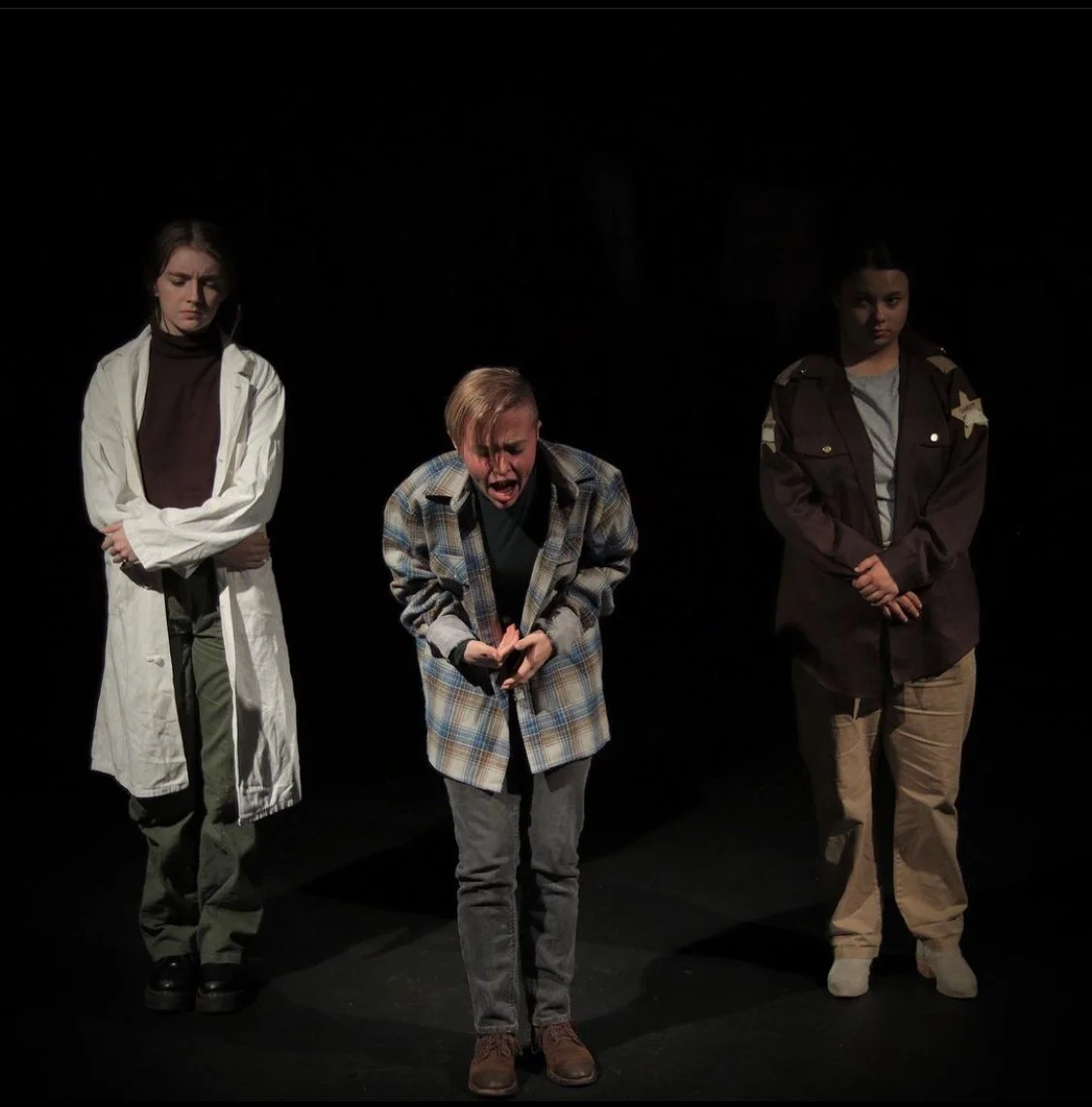 Three young girls on stage, one in the center with short blonde hair wearing a plaid jacket and expressing distress or sadness, flanked by two girls on either side with serious expressions; the girl on the left has long brown hair and wears a white c