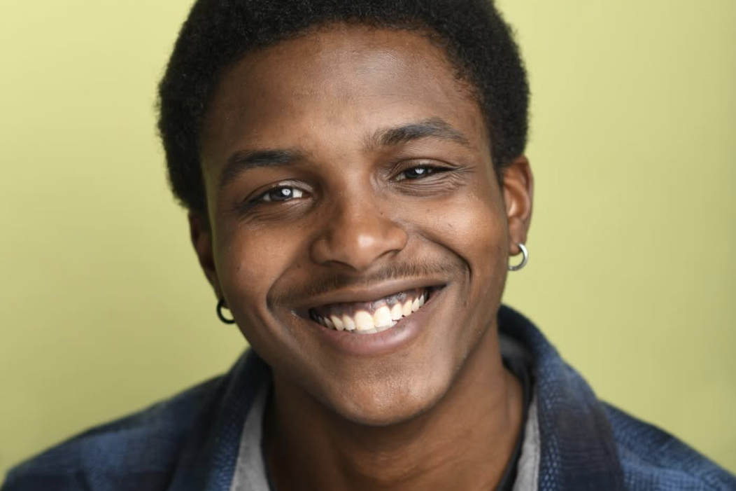 Close-up of a young man with a big smile, wearing hoop earrings, dark hair, and a plaid shirt, standing against a light green background.