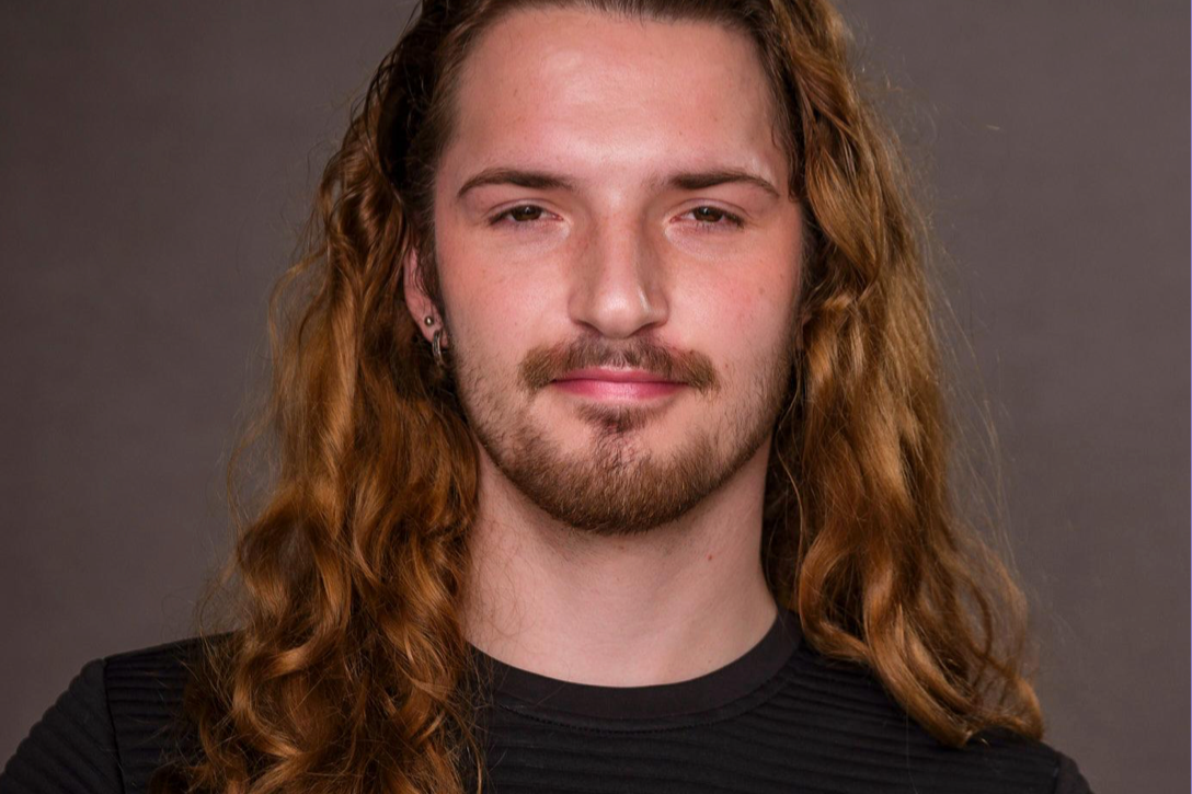 A young man with long curly red hair, a light beard, and earrings, looking at the camera with a neutral expression, wearing a black top against a plain background.