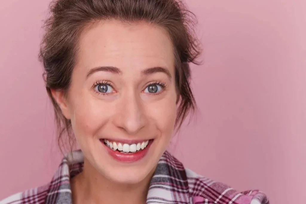 Portrait of a woman with light brown hair and blue eyes, smiling, wearing a checkered shirt, against a pink background.