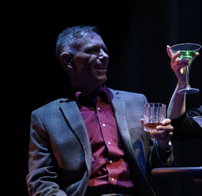 Man in a plaid blazer and red shirt smiling while holding a glass of whiskey, with a woman beside him holding a martini glass with a green drink.