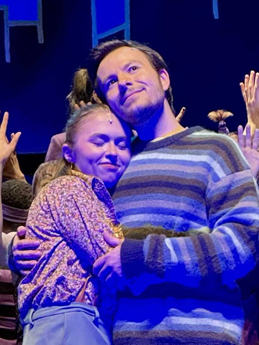 Two people hugging and smiling at a concert or event, with a dark background and blue lighting.