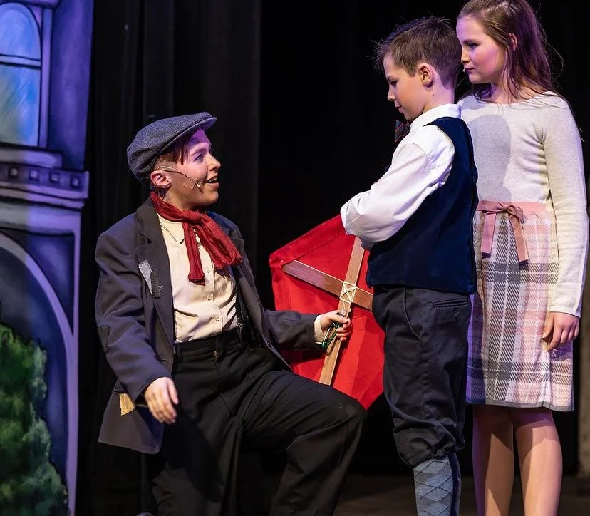 Three children on stage during a play, with one dressed as a character with a gray hat and red scarf, holding a sword, and the other two standing nearby.