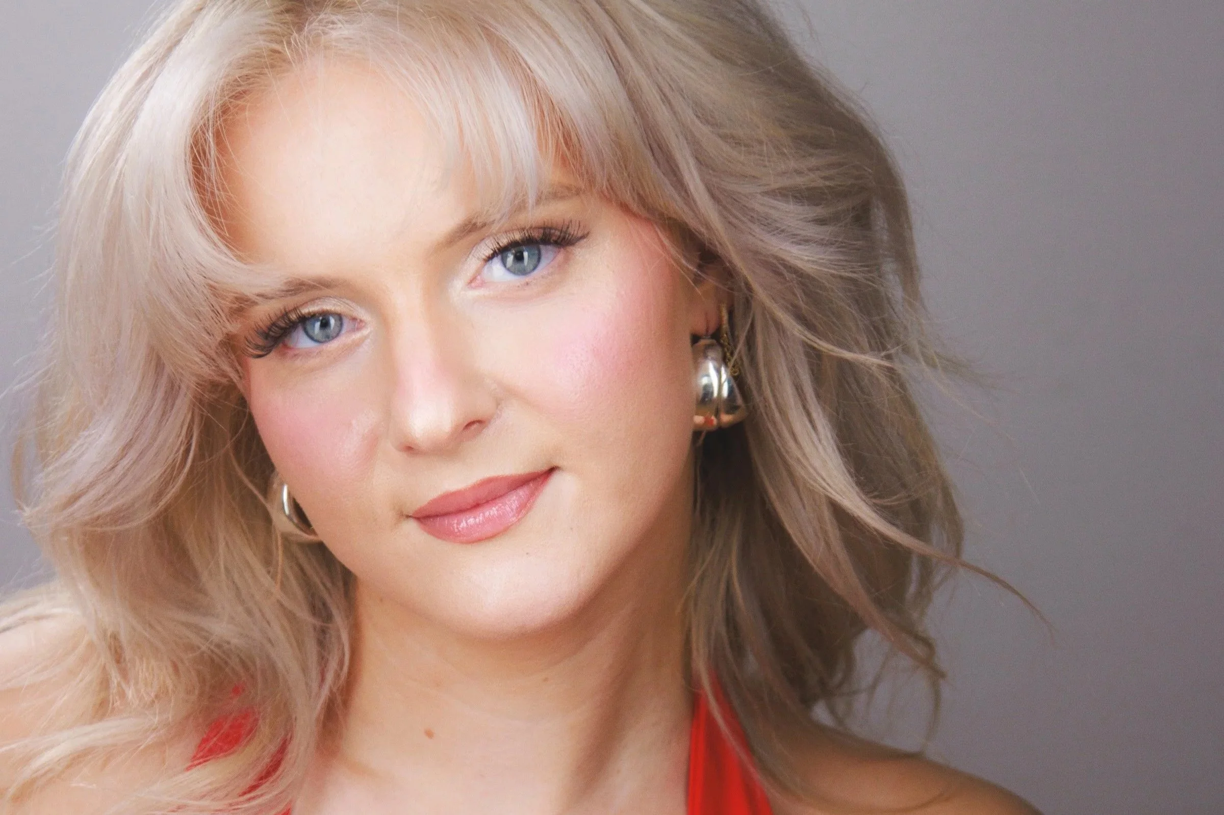Close-up of a smiling woman with blonde wavy hair, blue eyes, wearing earrings and a red top.