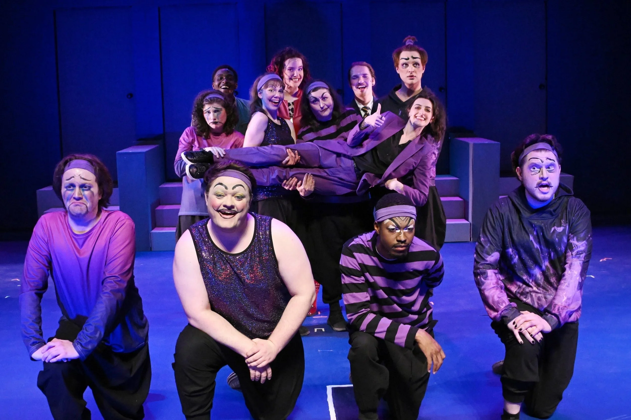 A group of actors in theatrical costumes and makeup pose on stage, with one man being carried by others, in front of a dark blue backdrop.