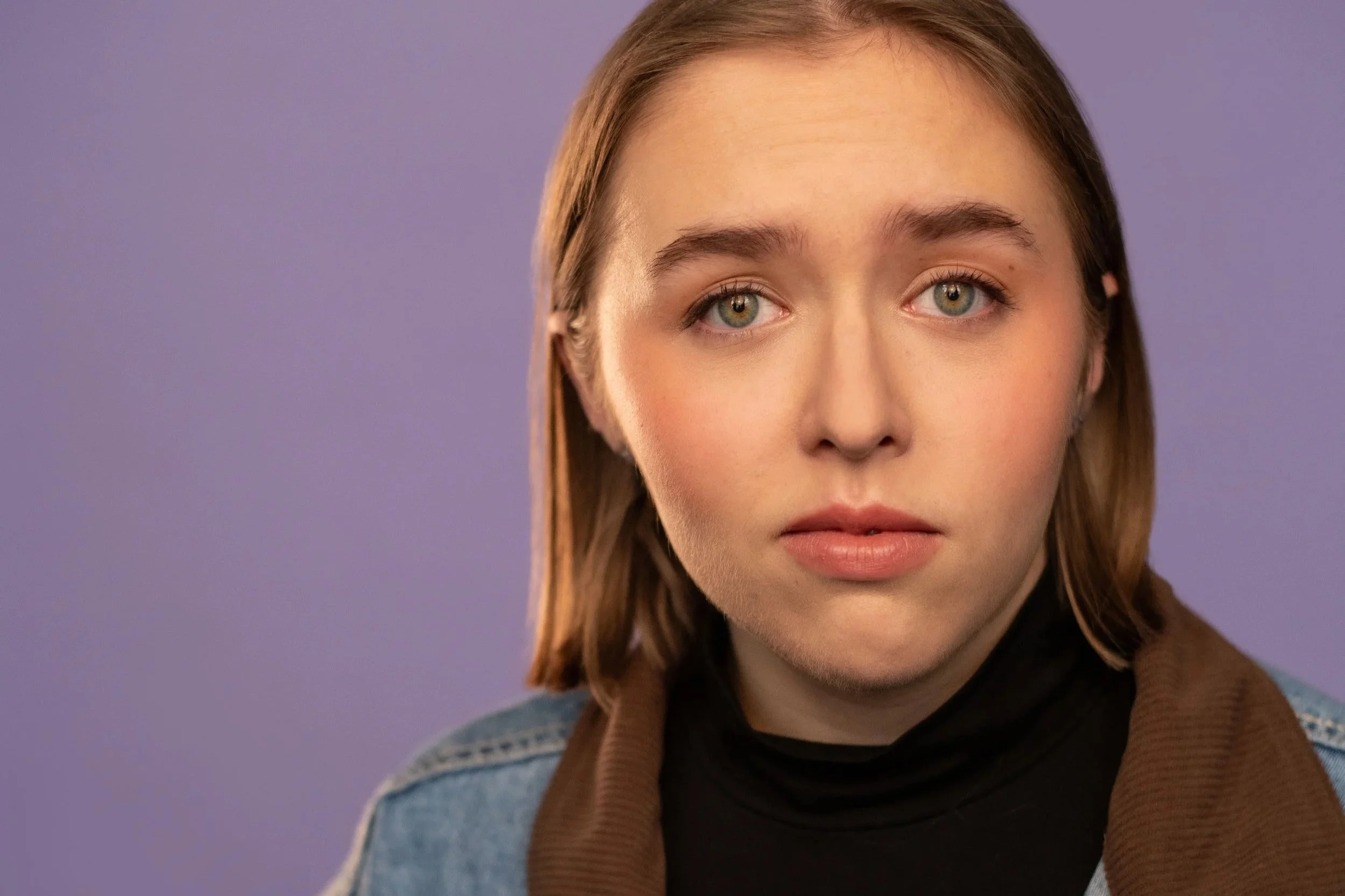 Close-up of a smiling young woman with light brown hair and blue eyes, wearing a white collared shirt and a red and yellow argyle sweater vest, against a pink background.