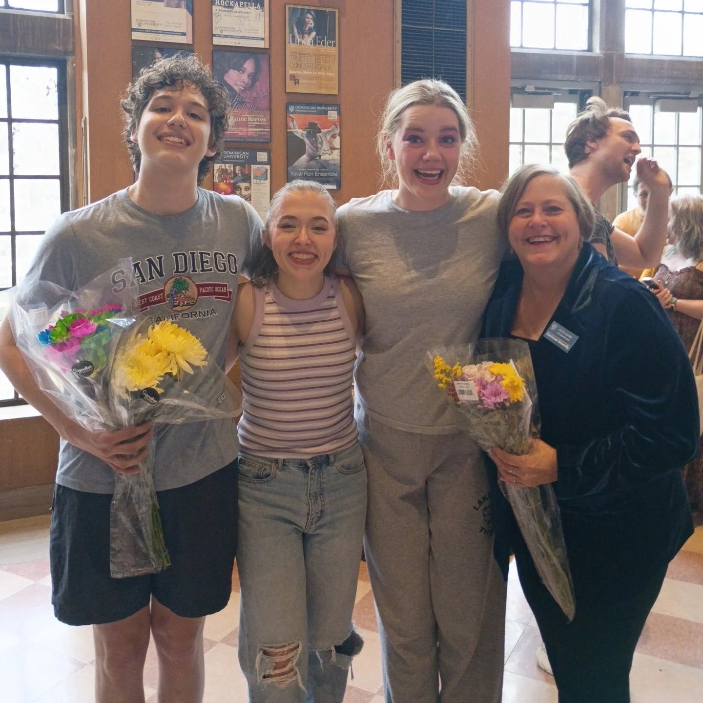 Four women and one man standing indoors, smiling and holding bouquets of flowers, celebrating together in a warmly lit room with wooden walls and posters on the background.