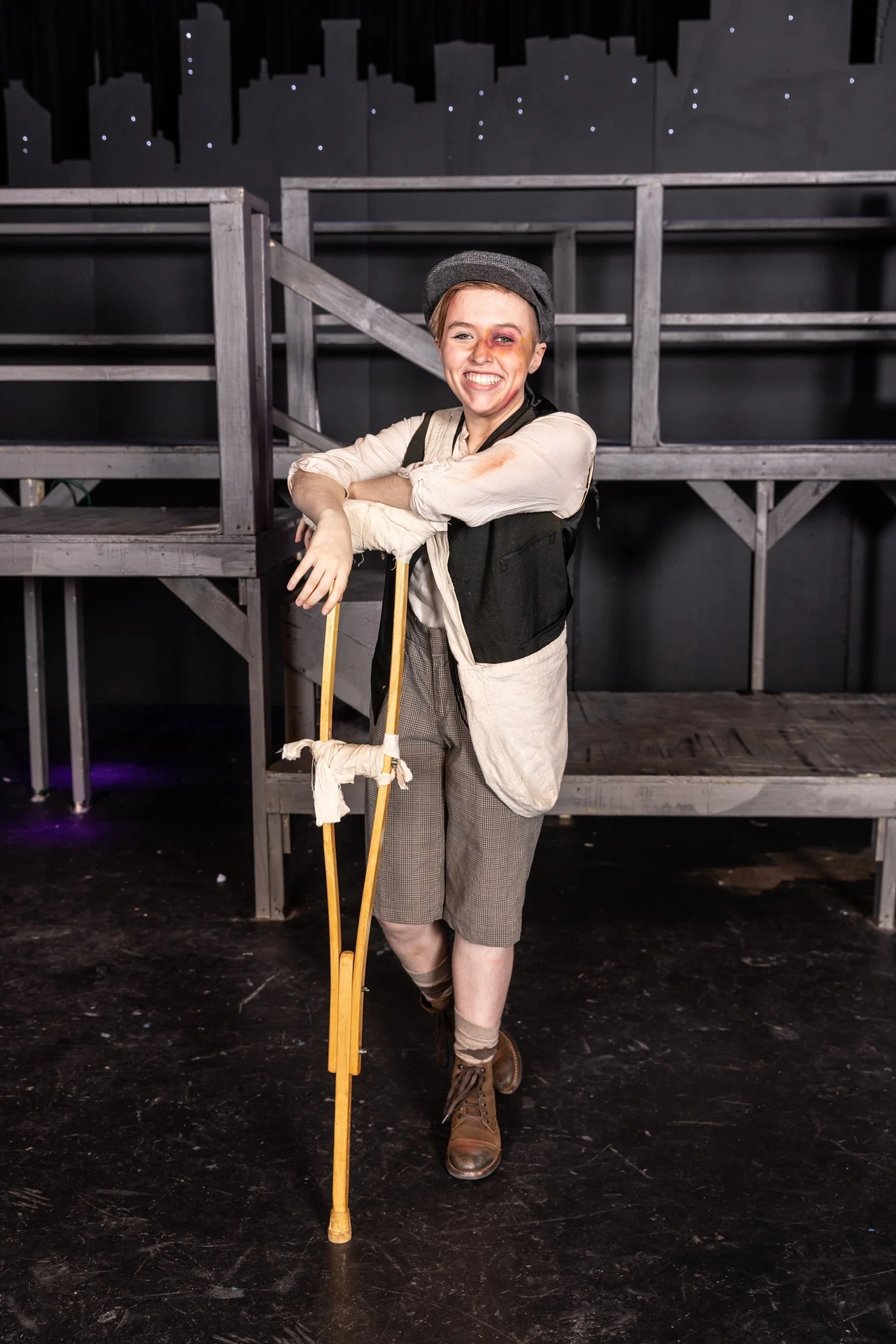 Person dressed as a theatrical character, with a bruised eye and smudged makeup, standing backstage with arms crossed and smile, holding crutches, in front of black stage set.