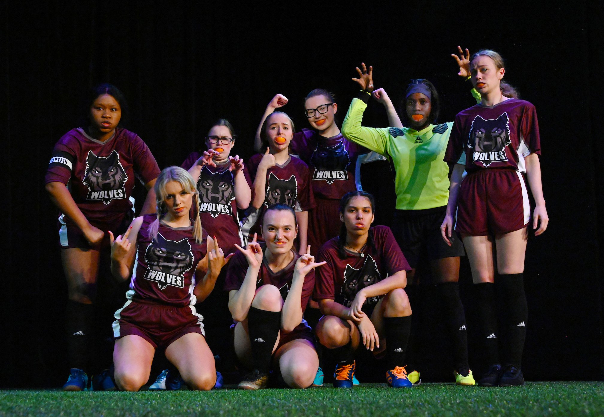 A group of young women in sports uniforms, some wearing glasses, posing on a sports field. They are making various gestures and facial expressions, with some sticking out their tongues. Most are wearing maroon uniforms with a 'Wolves' logo, and one p