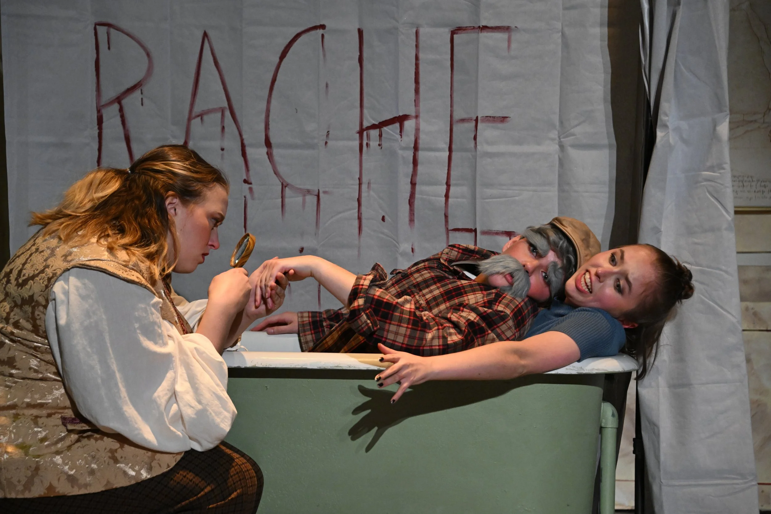 Two women in costume, one with gray beard and cap and the other smiling, lying on a table hand-in-hand, with a blood-smeared banner and a person examining the hand with a magnifying glass in the background.