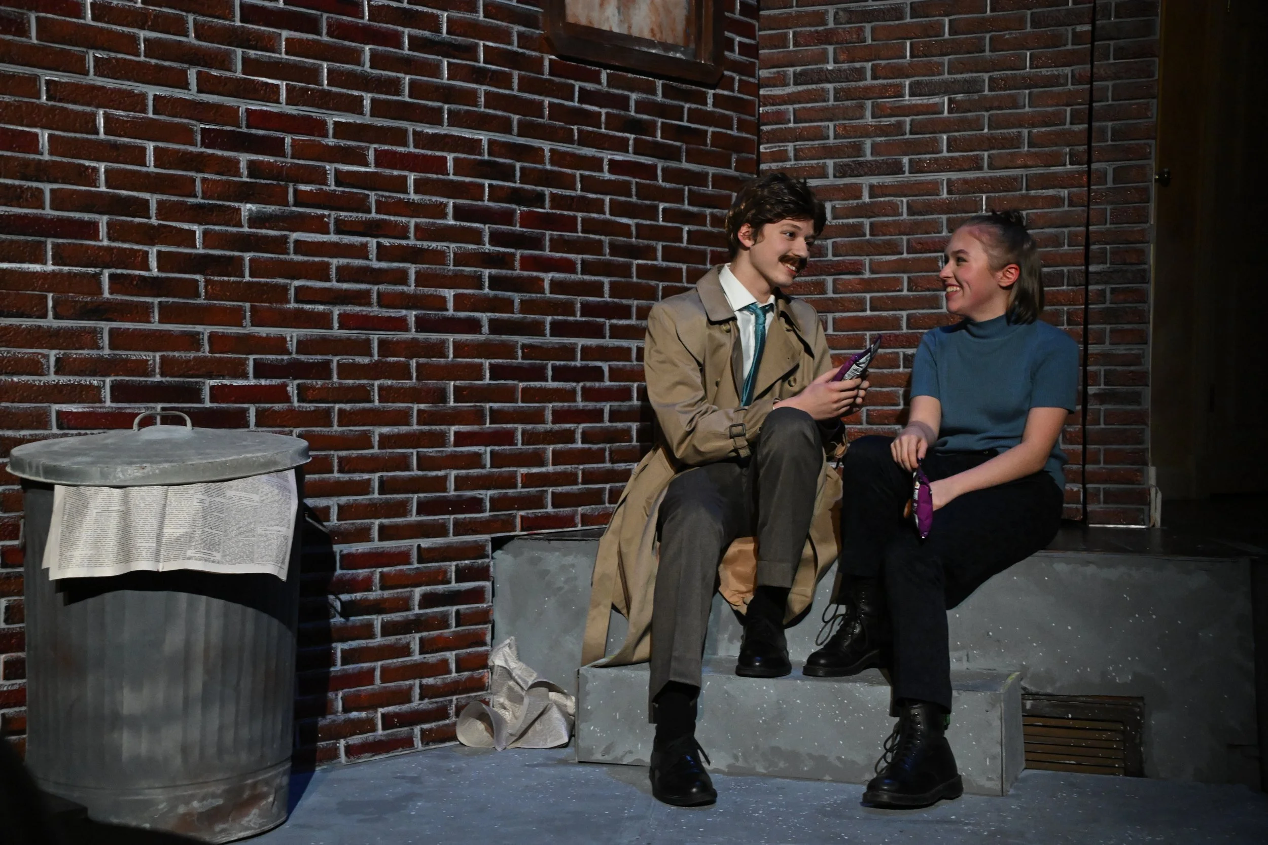 Two people sitting on a concrete block against a brick wall, smiling and talking. One is a man with a mustache and dark hair, holding a chocolate bar, and the other is a woman with dark hair tied back, wearing a blue shirt and black boots. There is a