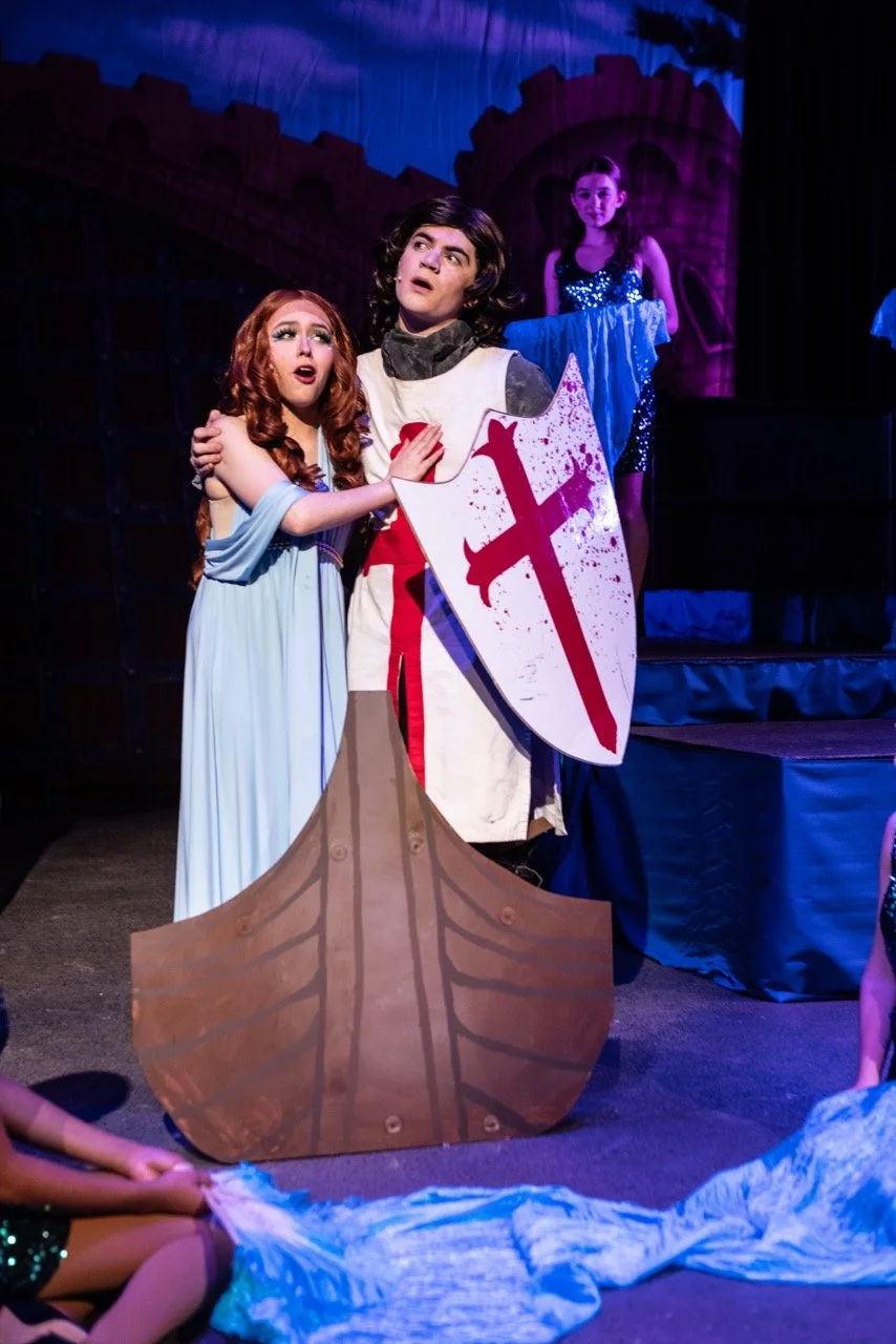 Three children in a theatrical play are dressed as medieval characters, with one holding a shield with a red sword symbol, on a stage with castle backdrop.