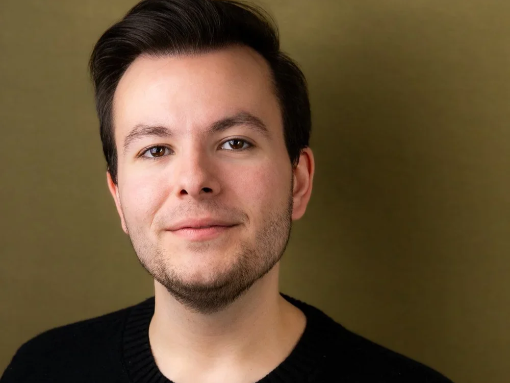Close-up portrait of a young man with fair skin, dark brown hair, and slight facial hair, wearing a black sweater against a neutral background.