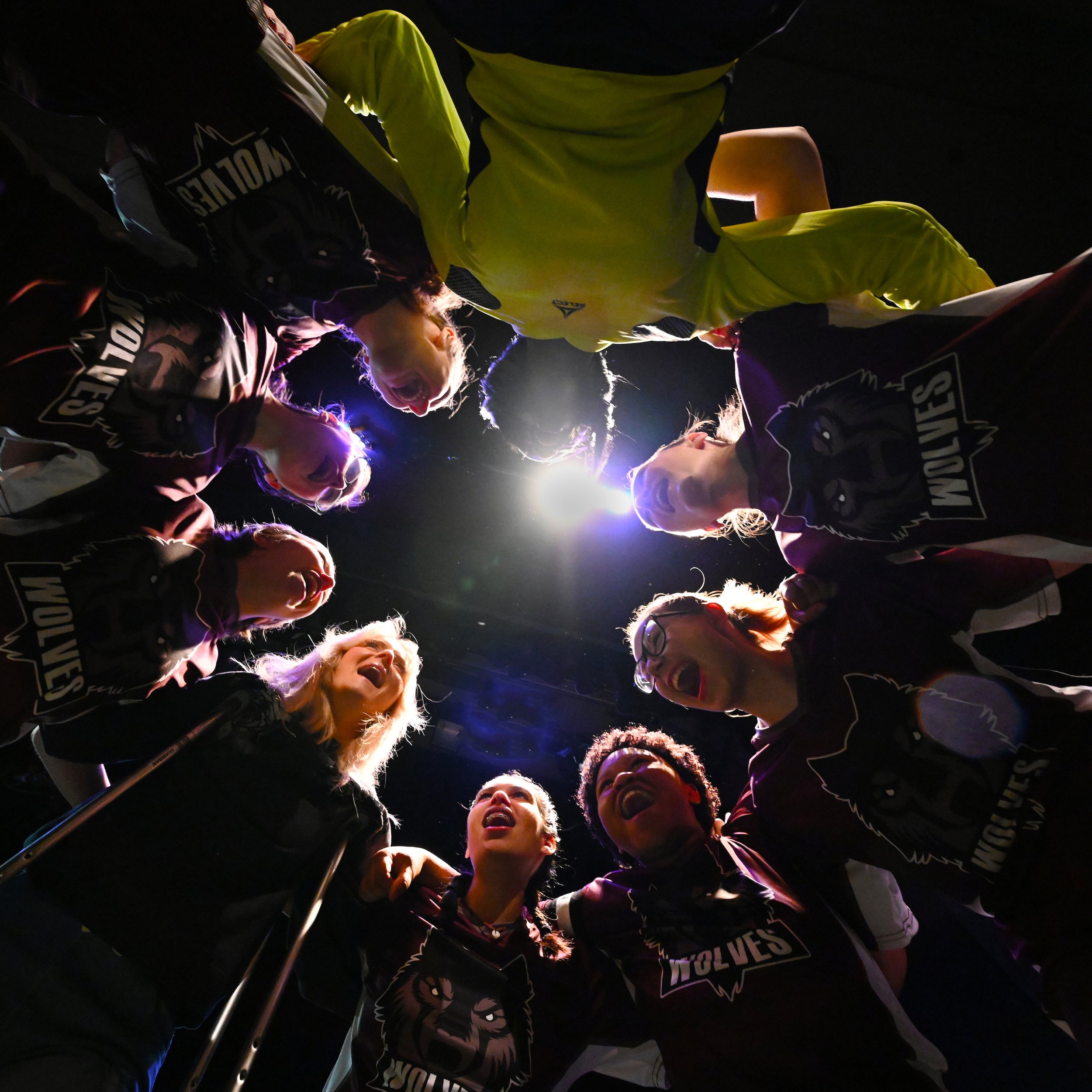 Group of athletes wearing sports uniforms and yellow jackets, standing in a circle and cheering or shouting, illuminated by bright lights in an indoor arena.
