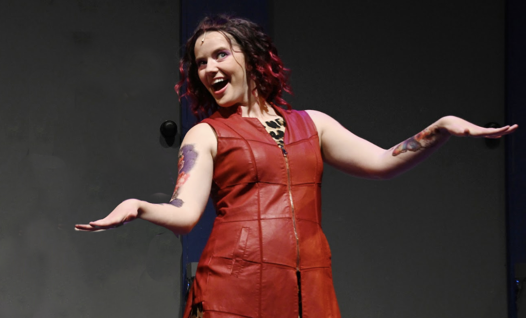 Woman with dark wavy hair and purple highlights, wearing a red leather dress, smiling and gesturing with her arms out to the sides.