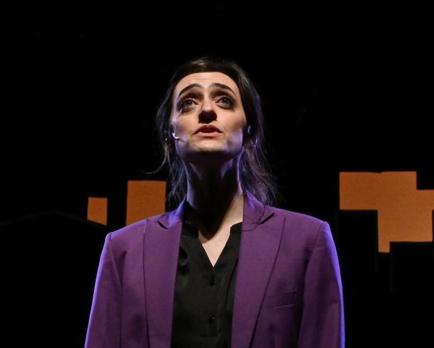 A woman with dark hair and makeup, wearing a purple blazer and black shirt, looks upward against a dark background with cityscape silhouettes.