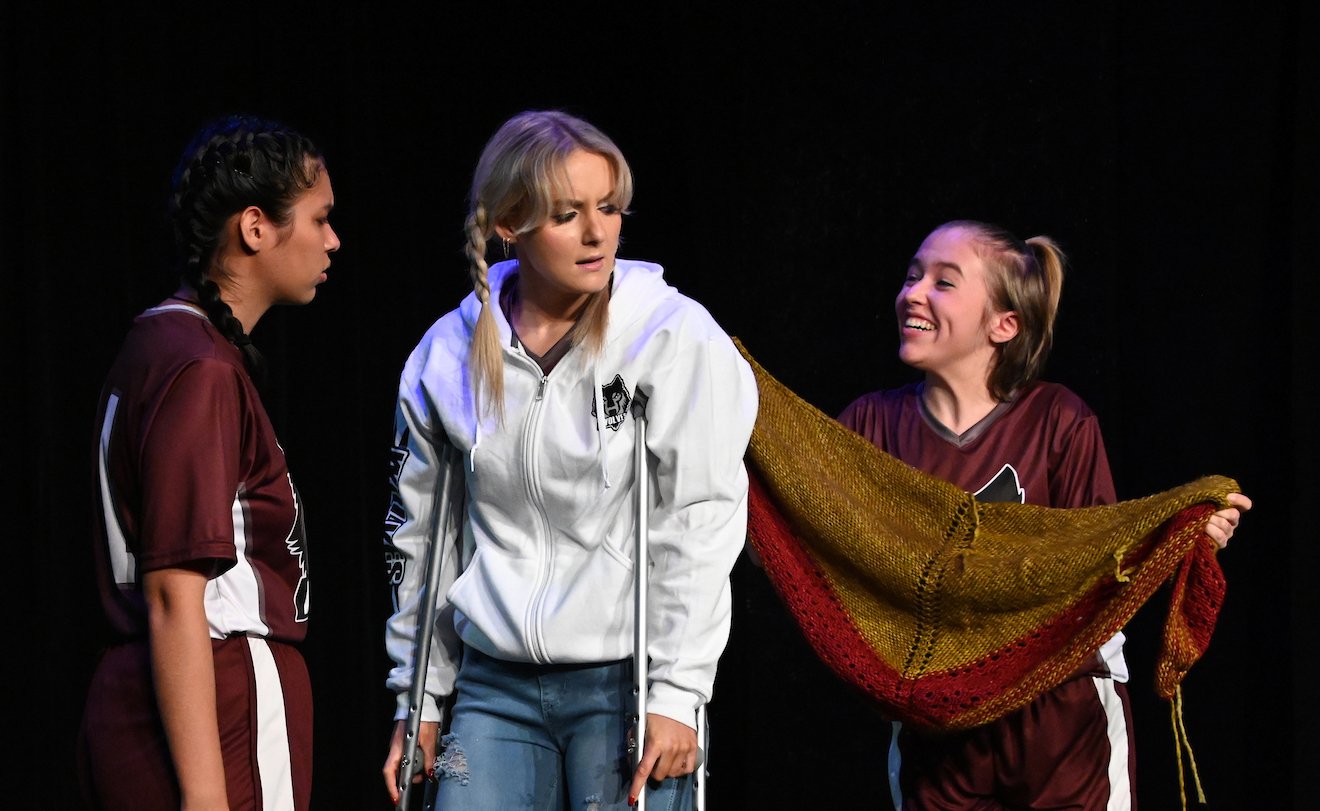 Three young women on stage, one in a white jacket with crutches, two in maroon jerseys, are engaged in a theatrical performance, with one holding a large, multicolored shawl.