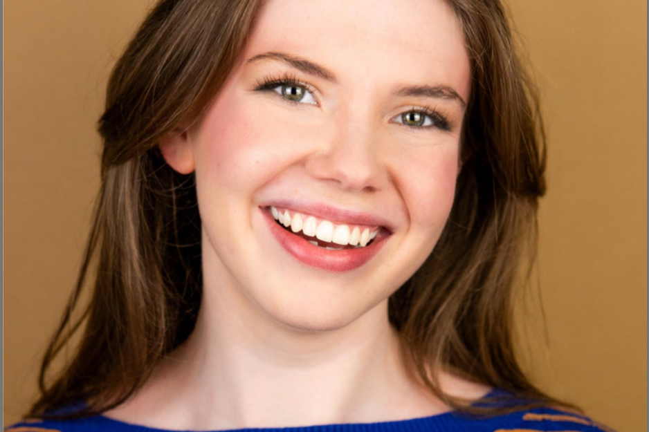 Close-up of a smiling woman with long brown hair, blue eyes, and wearing a blue top, against a brown background.