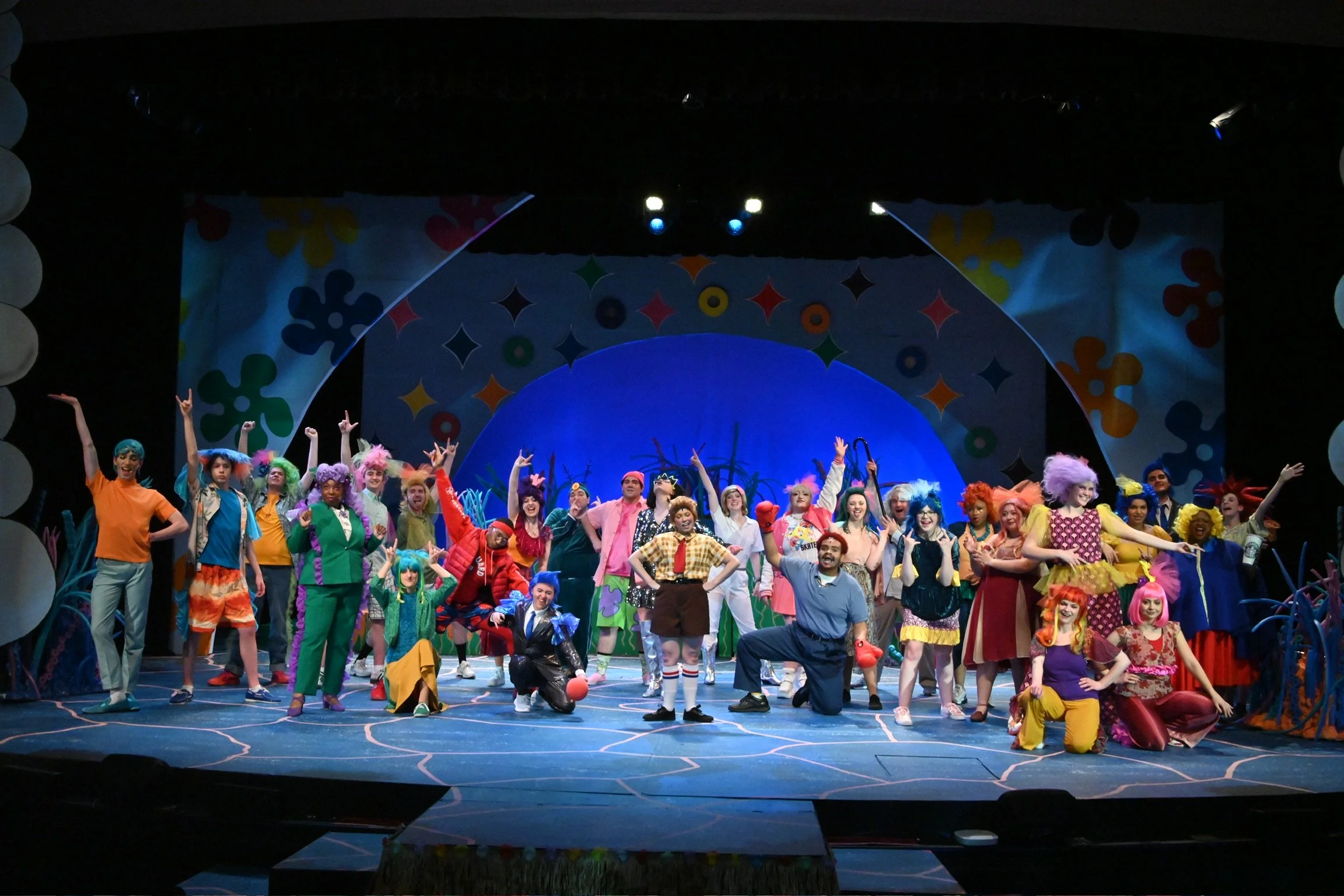 A large group of performers dressed in colorful costumes on stage, smiling and posing with some making gestures. The background features a vibrant, playful set with flower-shaped decorations in various colors.