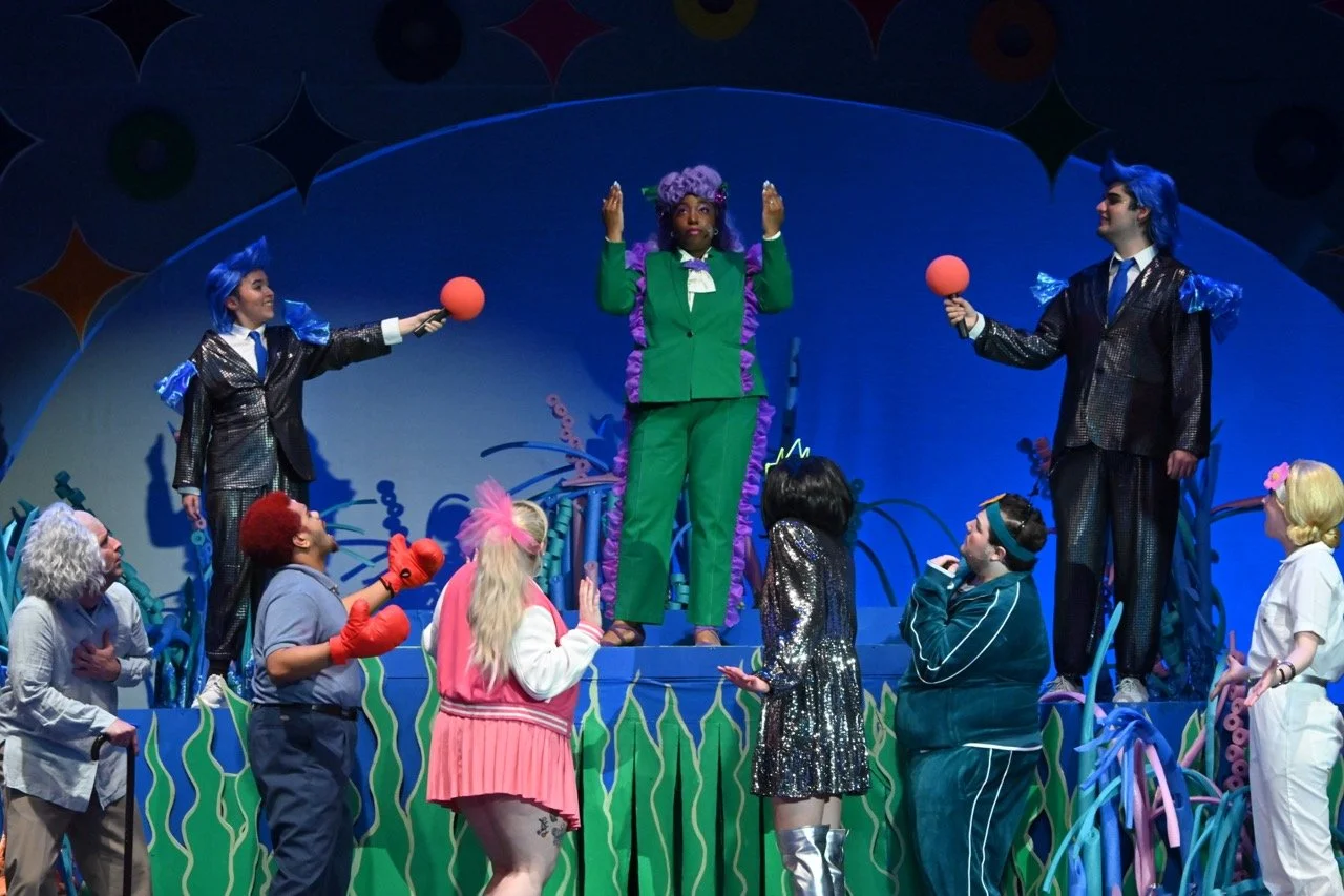 Theater scene with multiple actors dressed in colorful costumes on a decorated stage. The central figure, a woman in a green suit with purple accents, stands on an elevated platform with arms raised. Two actors with blue hair and shiny suits hold mic