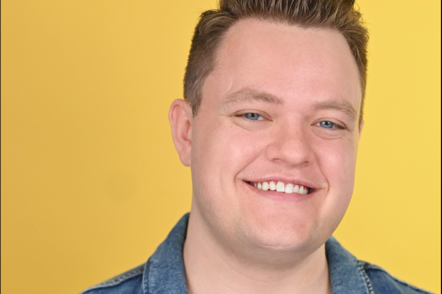 Close-up of a smiling young man with short light brown hair, blue eyes, wearing a denim jacket, against a yellow background.