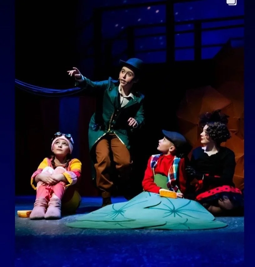 A theatrical scene with four characters, including a woman with black curly hair, two children in colorful outfits, and a man in a green jacket and hat, engaging in a storytelling performance on stage with a blue and orange set background.