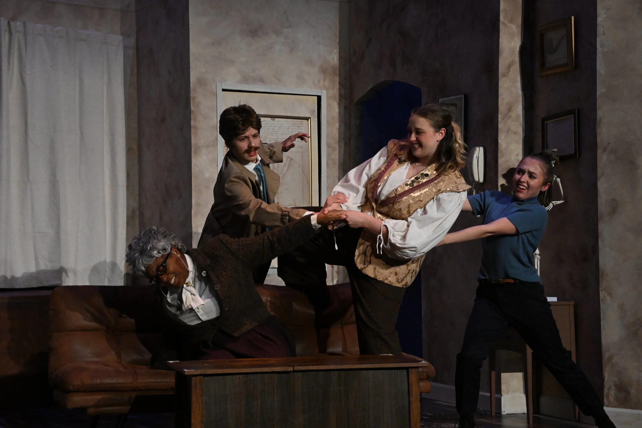 People acting in a theatrical scene, including a woman being held by others, a man with a mustache wearing a brown suit, an elderly woman with glasses, and a young girl. The scene appears to take place in a living room with framed pictures on the wal