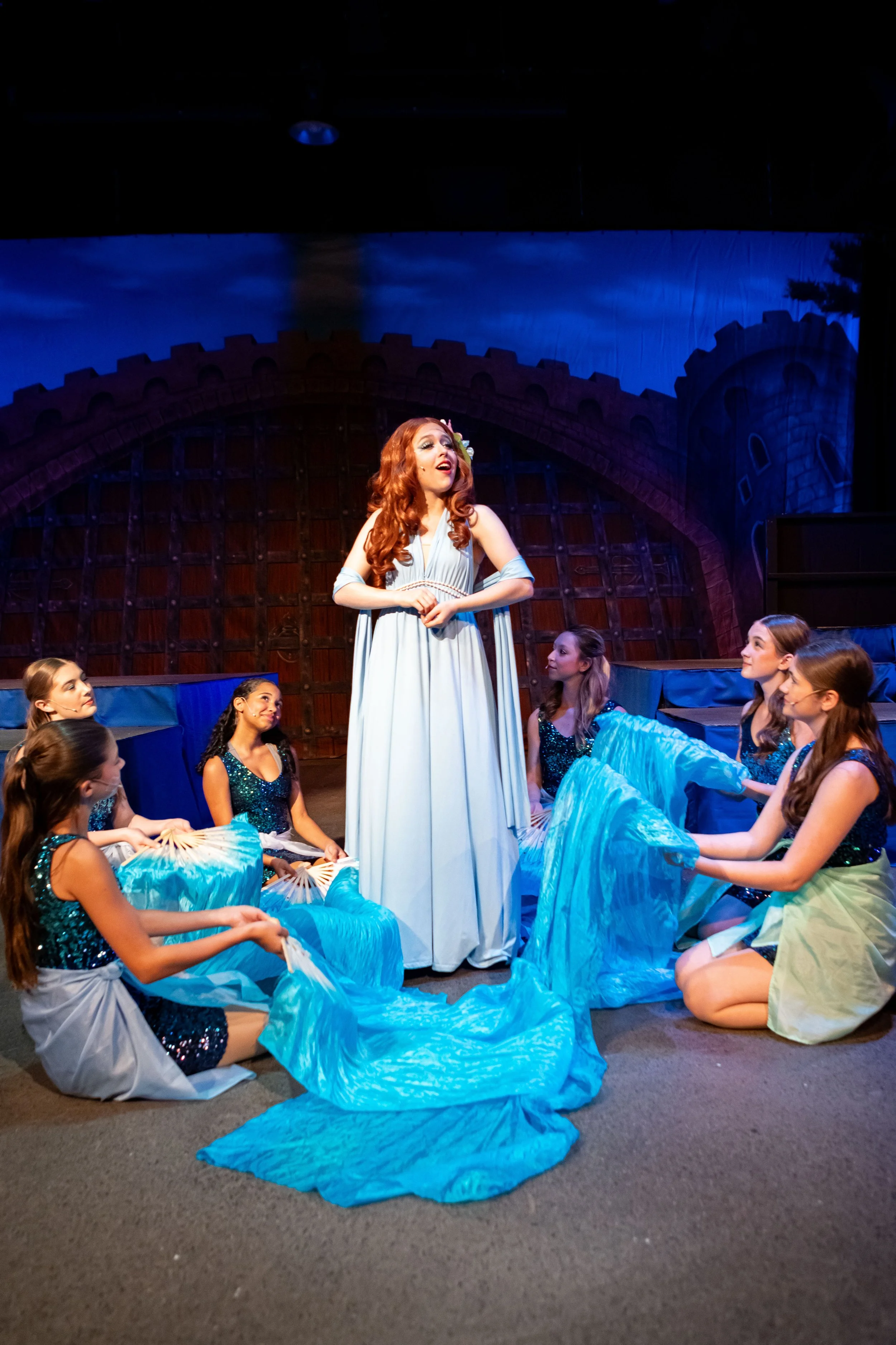 A theatrical scene featuring a woman with long red hair in a light-colored gown standing in the center, surrounded by young girls wearing matching sparkly blue and black dresses, sitting on the floor holding blue fabric. The stage background depicts 