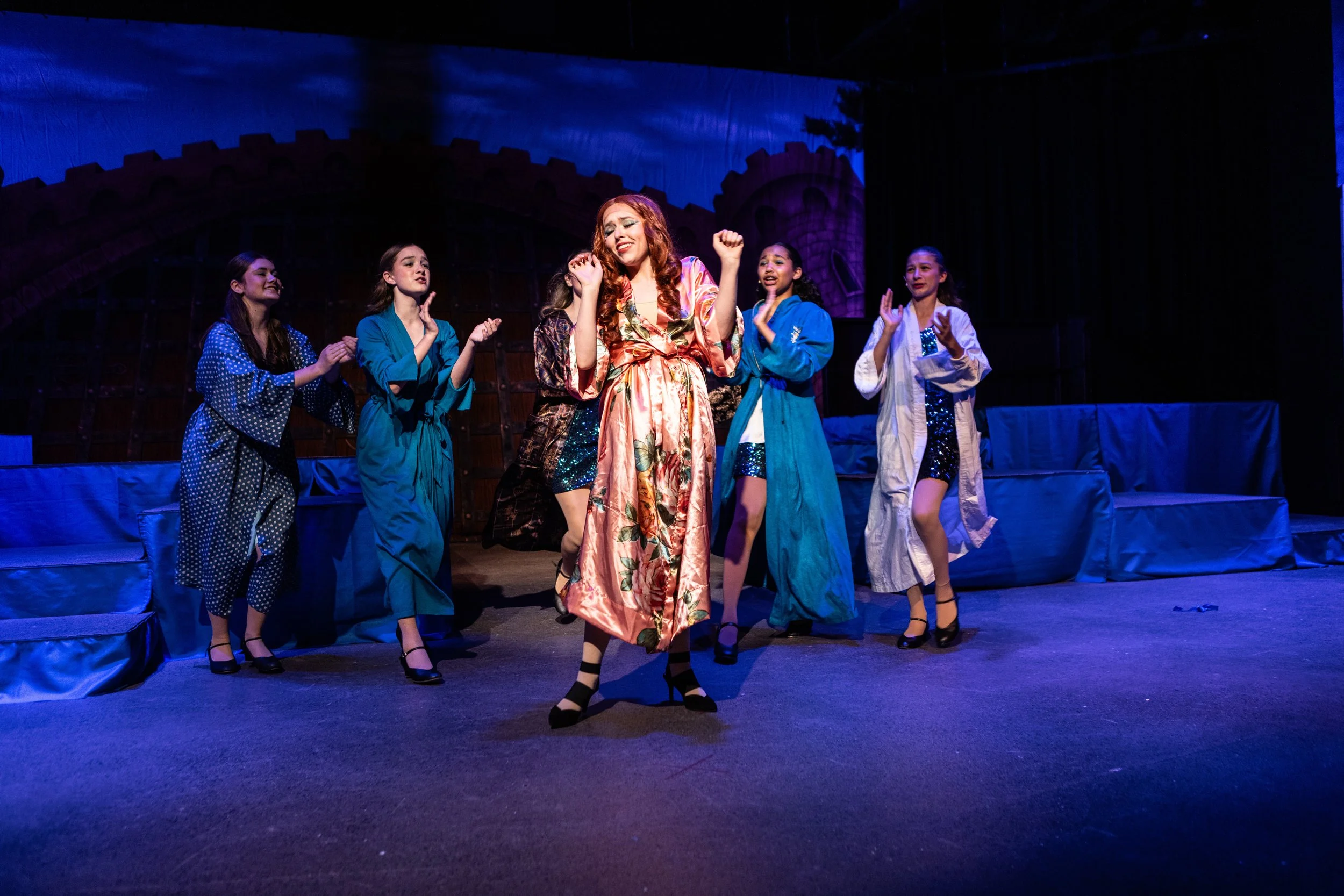 The image depicts a stage scene with six women dressed in colorful, patterned robes, performing a theatrical dance or musical. The woman in the center wears a floral satin robe, smiling and raising her arms, while the others, in blue and white robes,