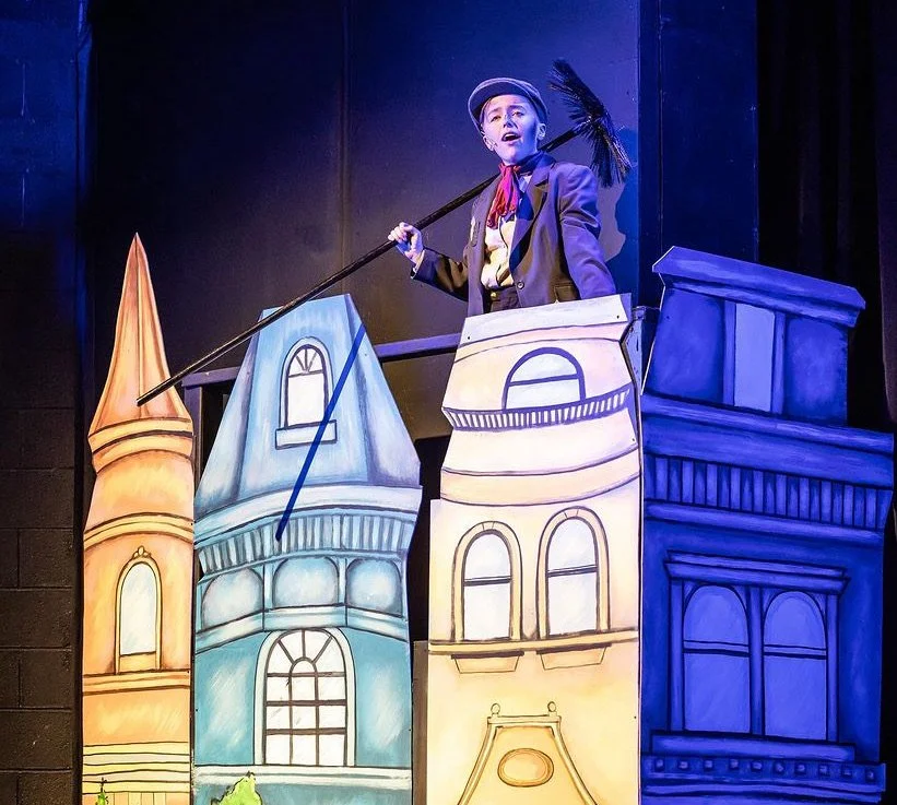 Child actor performing on stage in a play with colorful cardboard cityscape background, dressed in costume with a hat, blazer, and holding a broom.