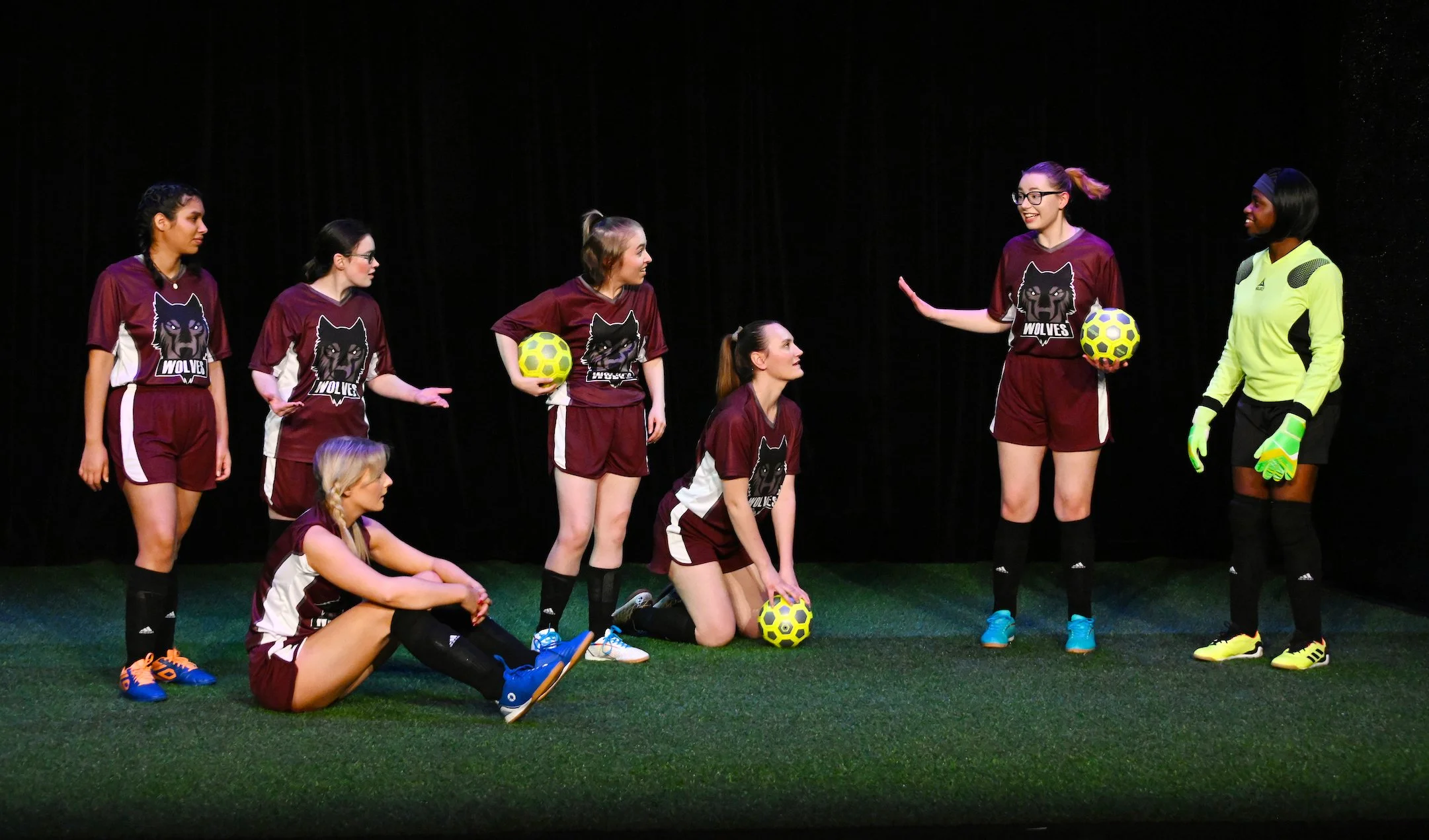 Group of female soccer players on stage, some sitting and some standing, wearing maroon jerseys with a wolf logo, holding soccer balls, with a black curtain background.