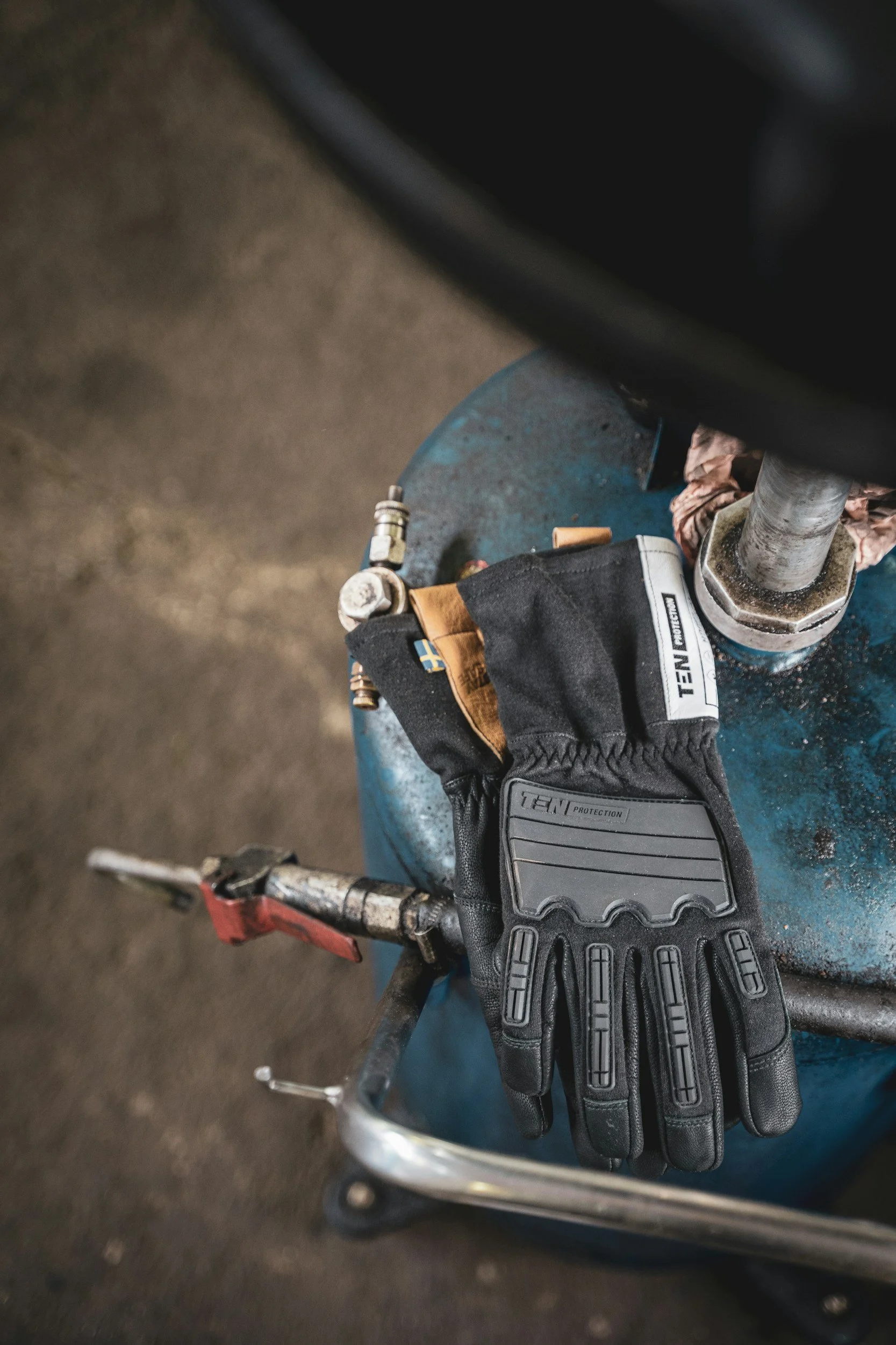Close-up of a person wearing black gloves with gray accents, holding a metal part, possibly a tool, on a blue work surface with various tools and equipment in the background.