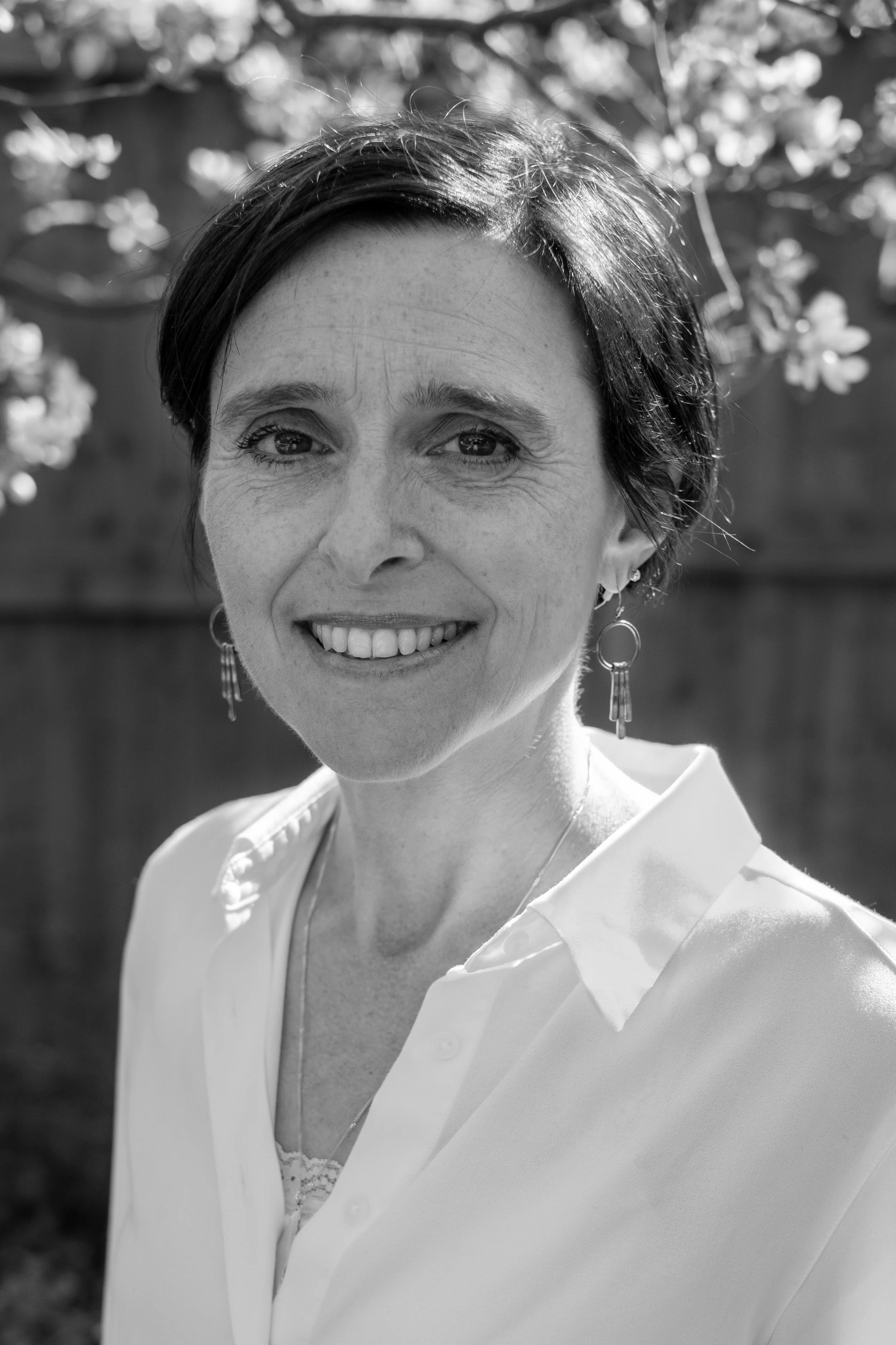Black and white close-up photograph of a woman smiling outdoors, wearing a white shirt with a partly open collar, earrings, and a necklace, with a blurred background of trees and flowers.
