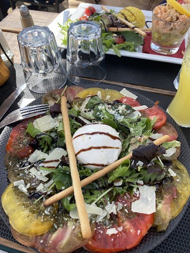 Salade avec tomate, mozzarella, feuilles vertes, olives, et gressins, accompagnée d'une boisson gazeuse, à côté d'autres plats et desserts.