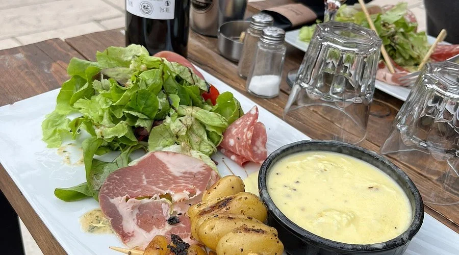Plate de charcuterie avec salade verte, pommes de terre rôties, andue au fromage, avec plusieurs verres et bouteilles de vin sur une table en bois.