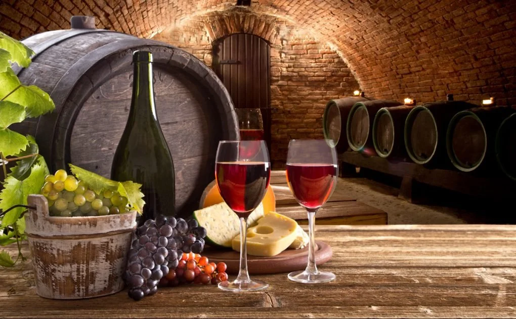 Verre de vin rouge, bouteilles de vin, fromages, raisins, et barriques dans une cave à vin en pierre.
