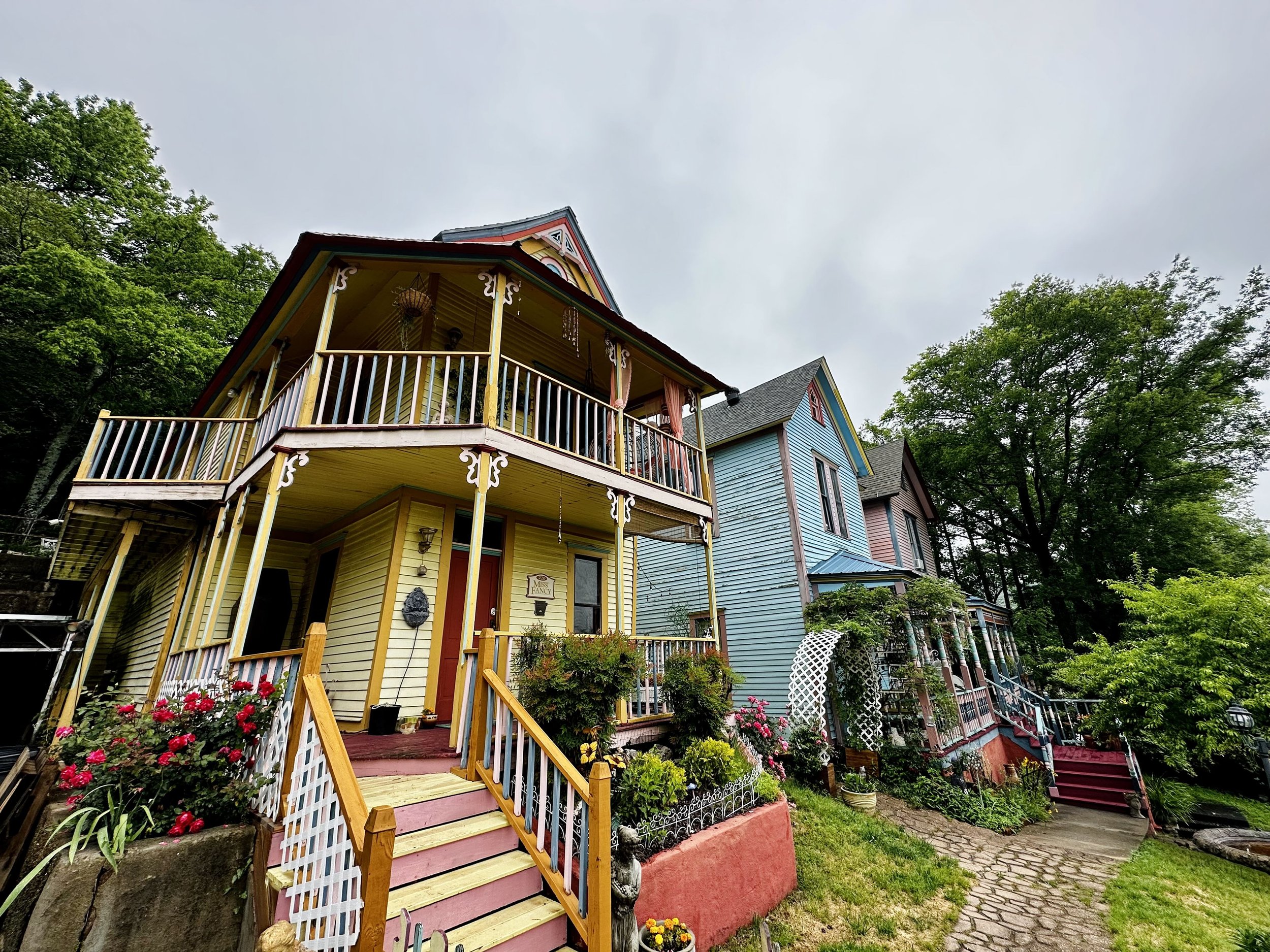 Colorful Victorian-style houses with porches and gardens, surrounded by trees under a cloudy sky.