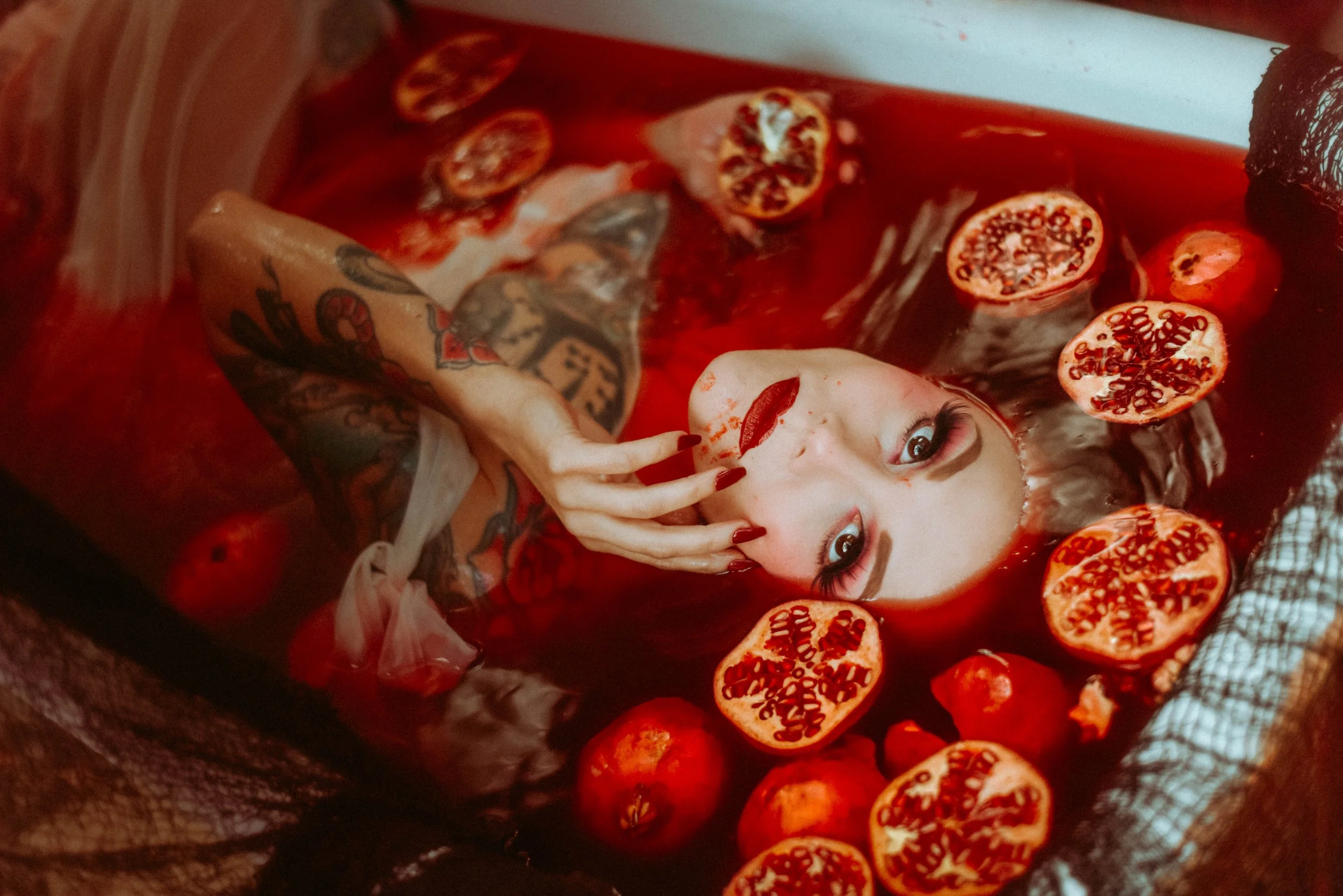 A woman with tattoos and dark lipstick submerged in a red liquid with sliced pomegranates floating around her, and looking up at the camera. A luscious and intimate look inside of our bathtub photoshoots.