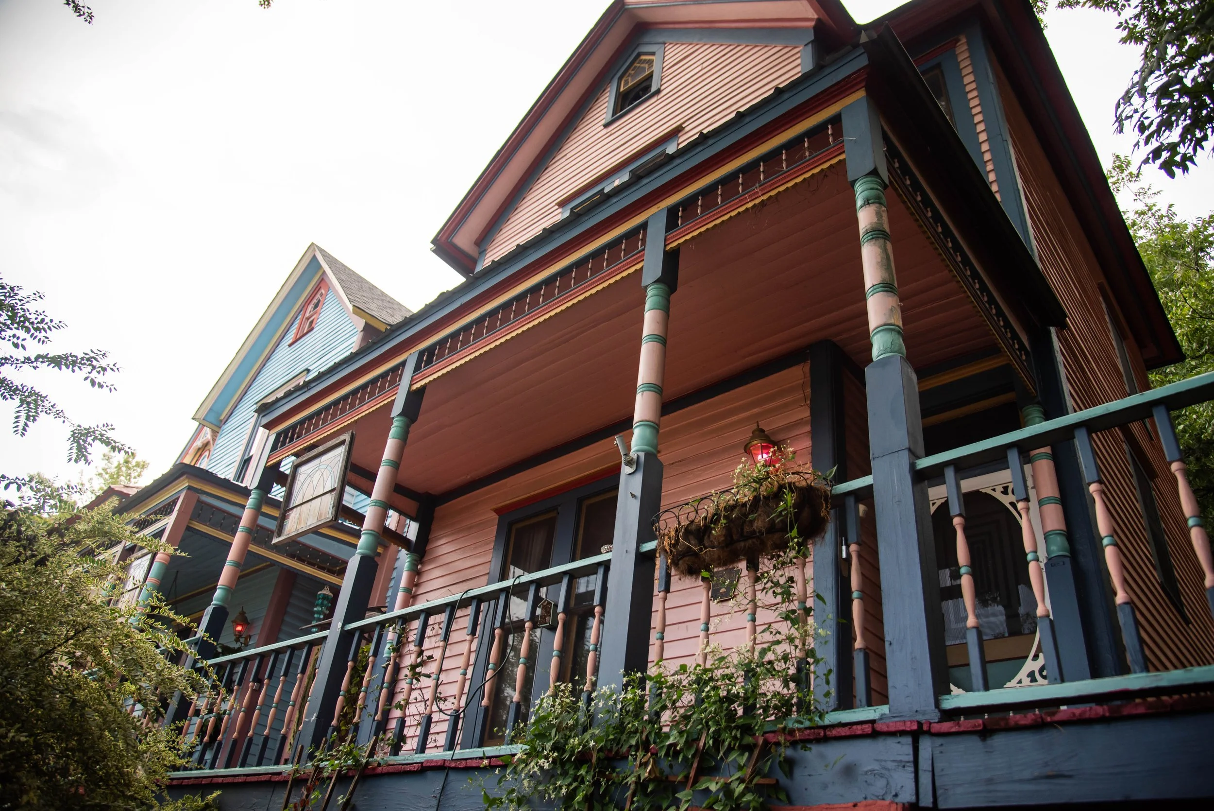 Colorful Victorian-style house with pink, blue, and yellow accents, surrounded by greenery and trees.