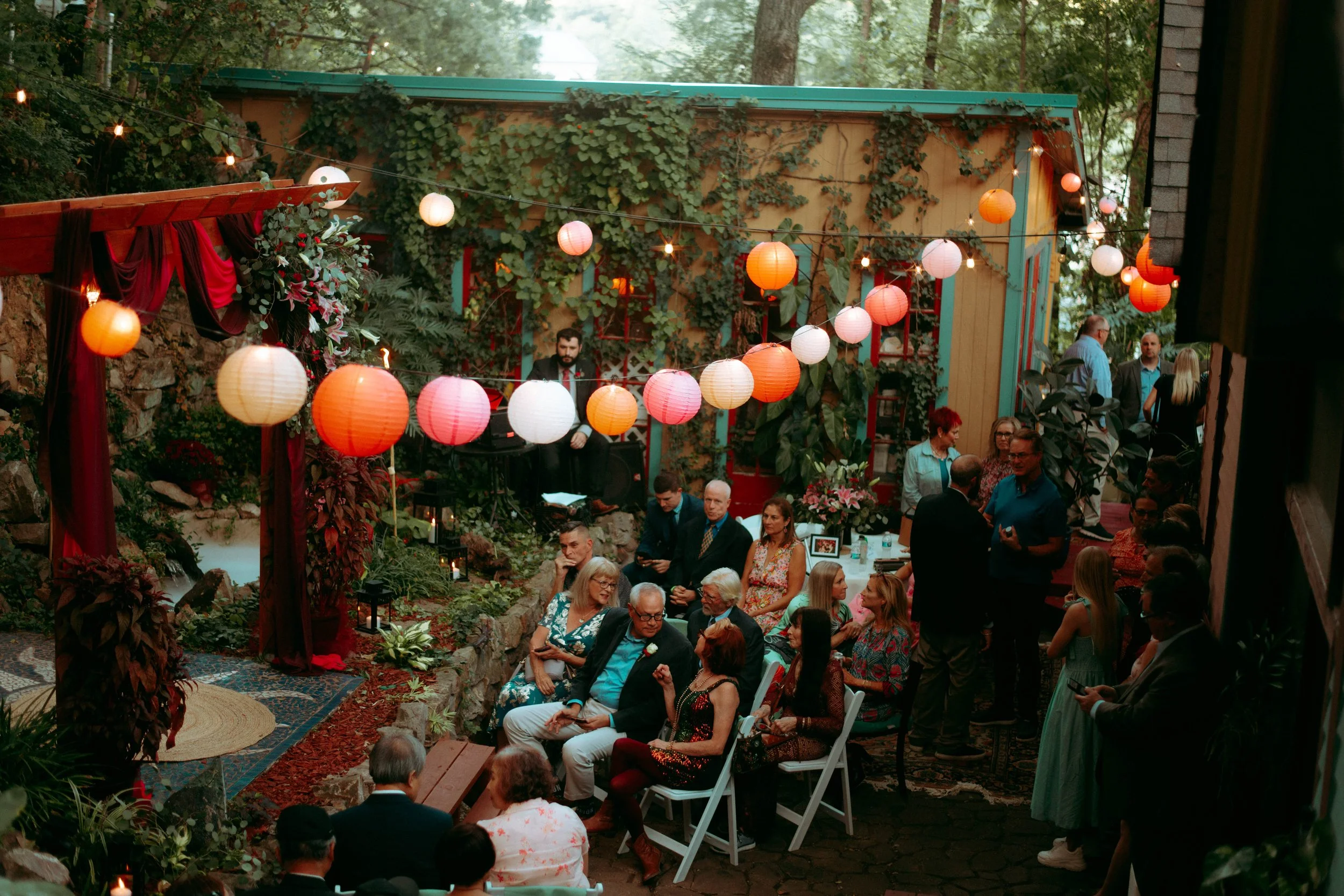 People gathered outdoors at a decorated venue with hanging paper lanterns and lush greenery, attending a celebration or event in the evening.
