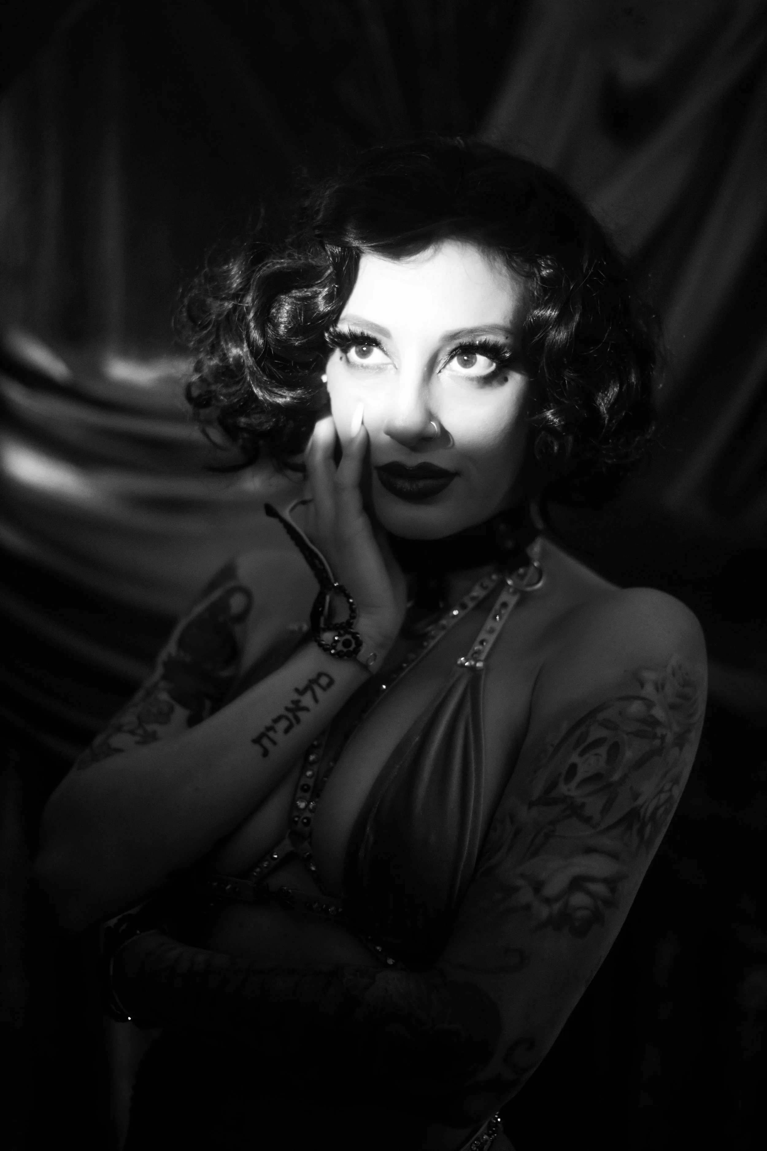 A woman with dark, curly hair and tattoos on her arms, wearing makeup and a nose ring, posing with her fingers near her face, in a black and white photograph.