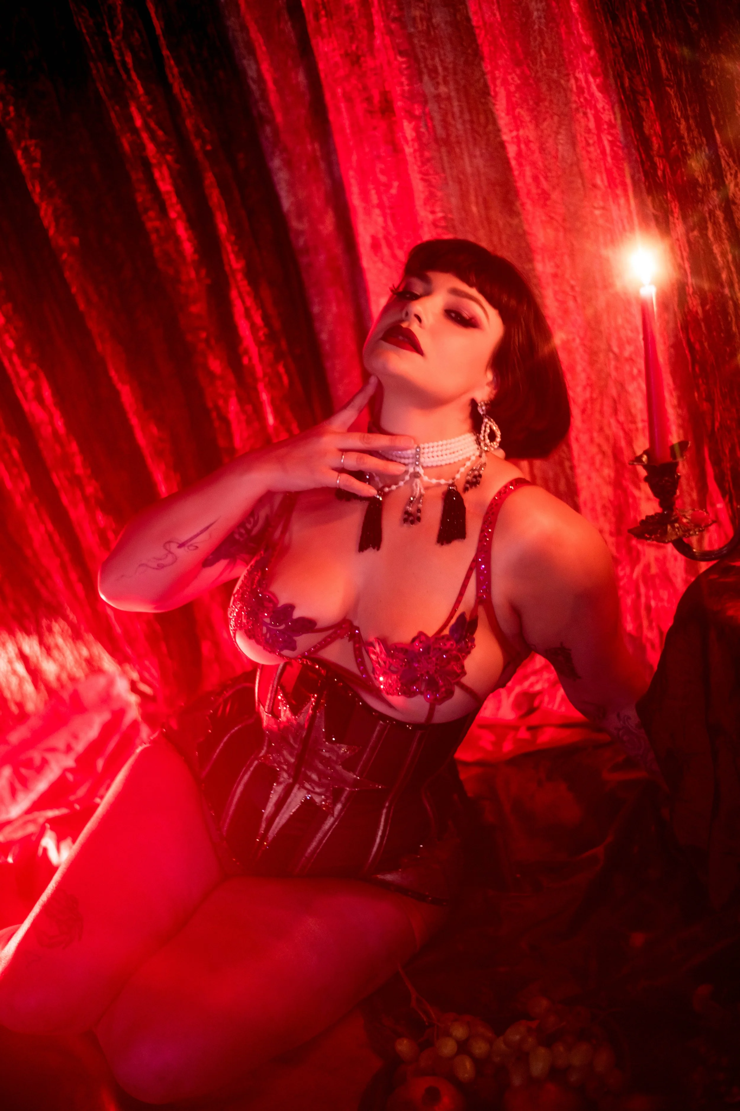 A woman with dark hair and makeup, dressed in a red and black lingerie outfit, posed with her head tilted and hand touching her chin, in a red-lit setting with a candle and grapes as props.