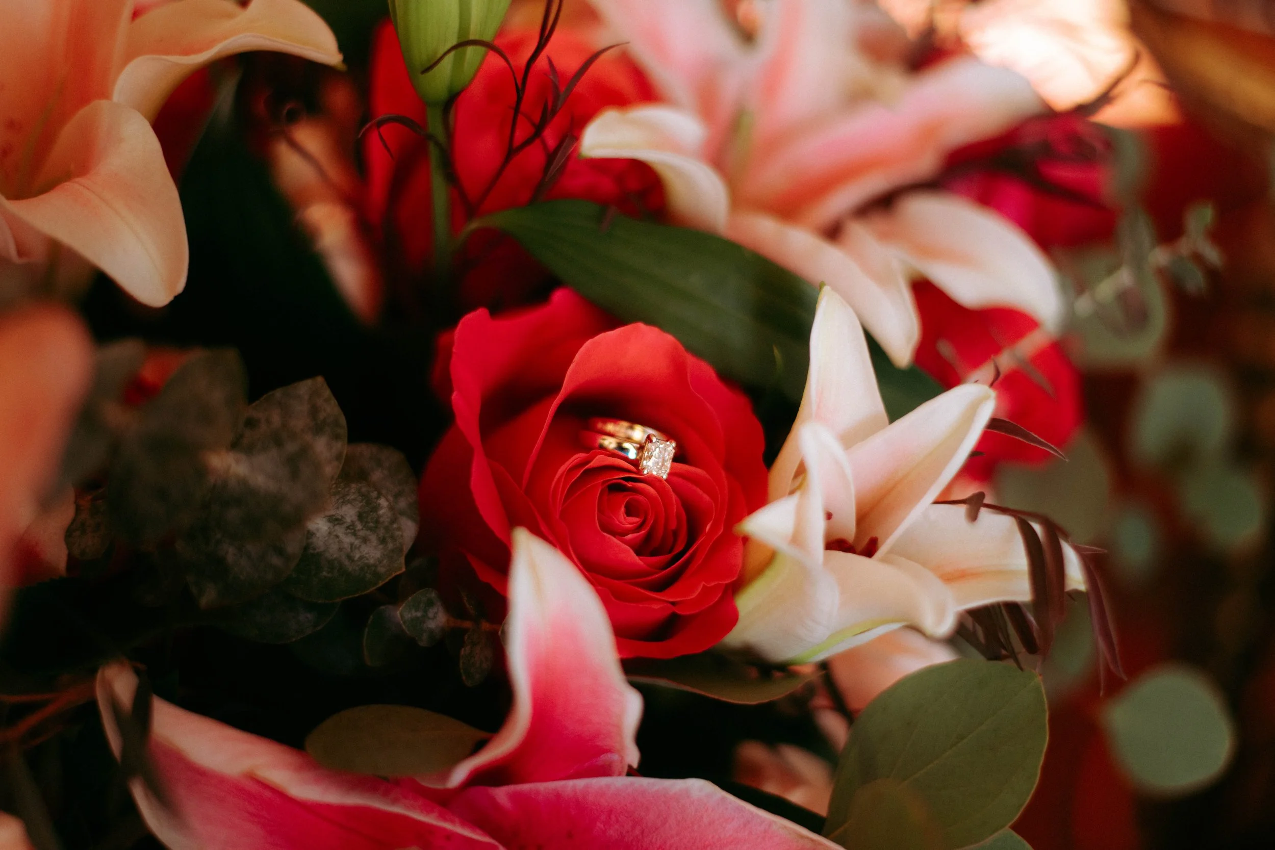 Close-up of a bouquet featuring a red rose with a diamond ring on its petals, surrounded by white lilies, pink flowers, green leaves, and dark foliage.