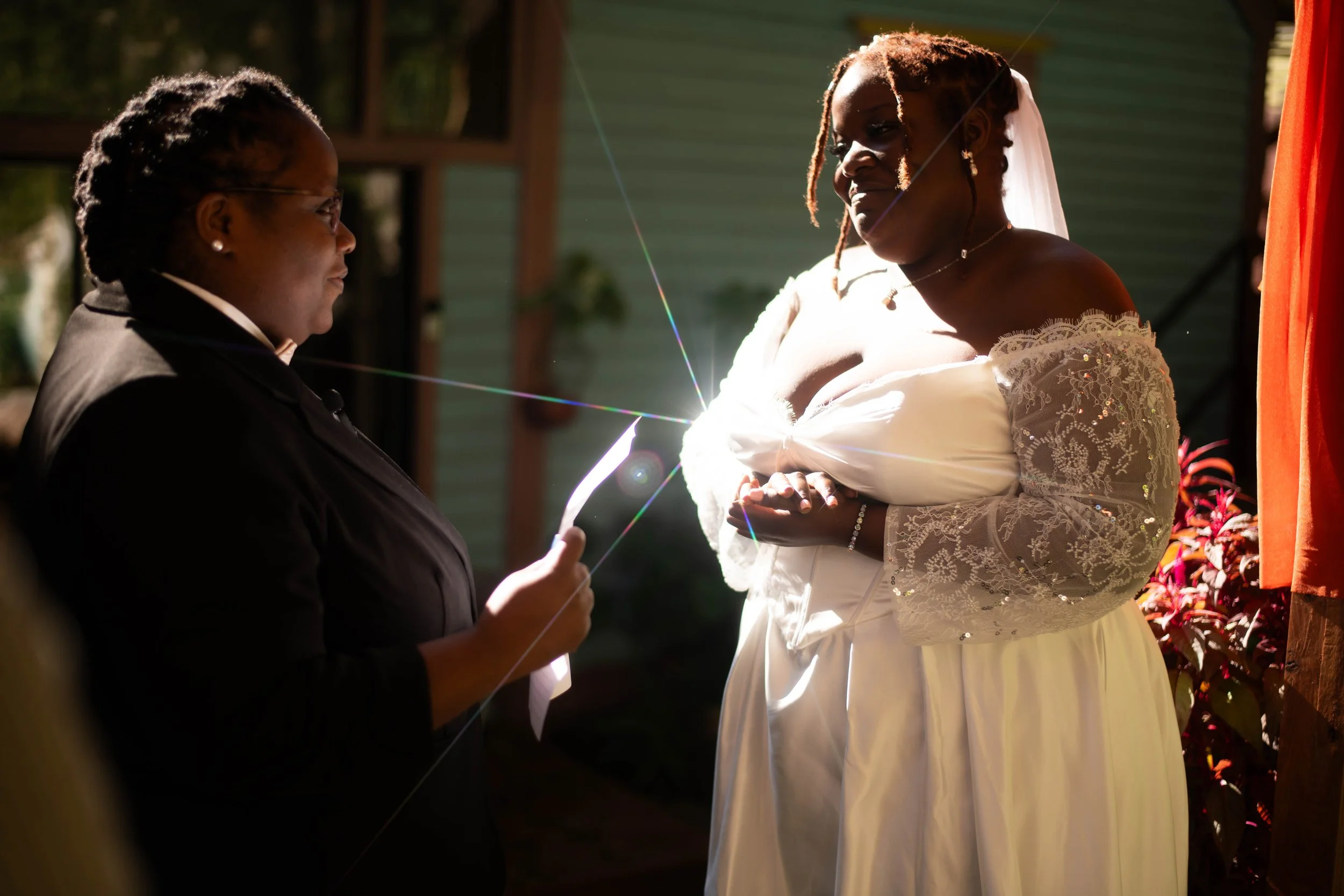 A woman in a white wedding dress with lace sleeves and a veil is standing outdoors, holding the hands of a man in a dark suit, during a wedding ceremony.