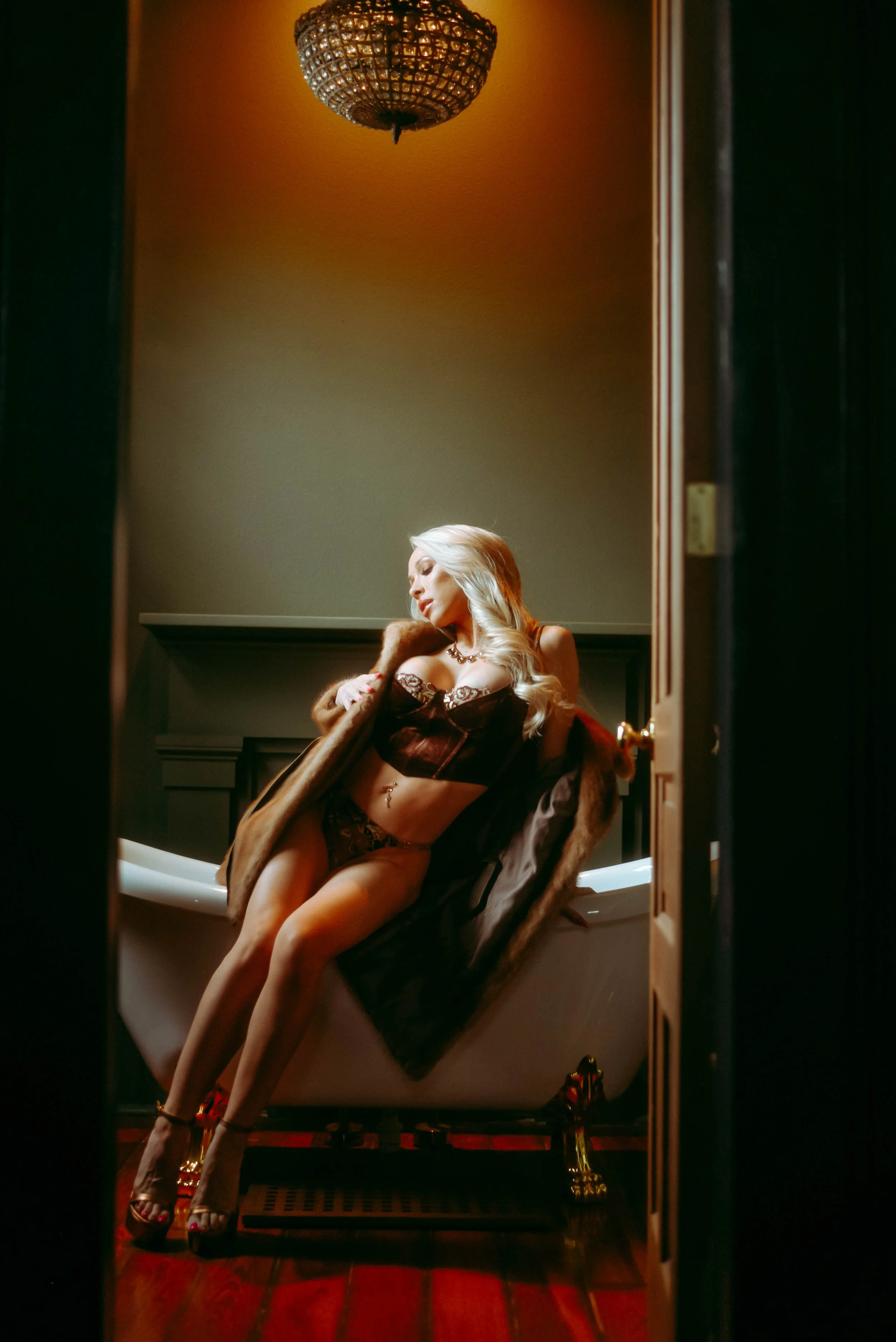 A woman with long blonde hair in lingerie and high heels, sitting on the edge of a bathtub in a dimly lit room, with a fur coat draped over her shoulders.