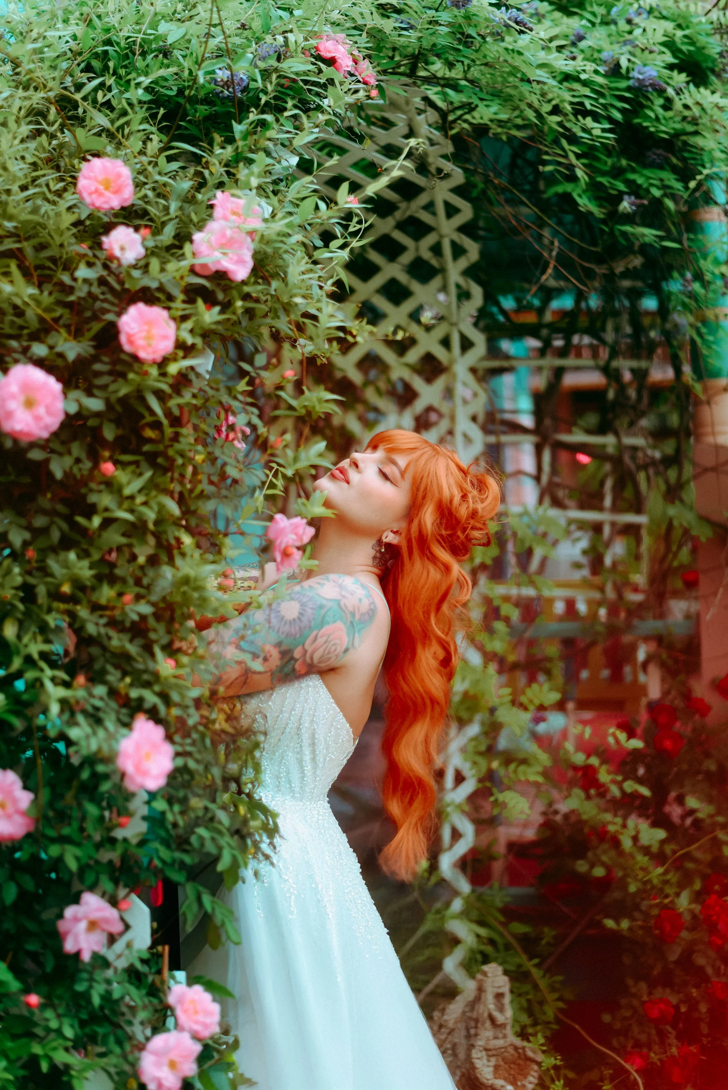 A woman with long, wavy red hair and a floral tattoo on her arm is smiling with closed eyes, leaning against pink flowers and green foliage in a garden setting. She is wearing a white, strapless dress and is surrounded by lush greenery and garden str