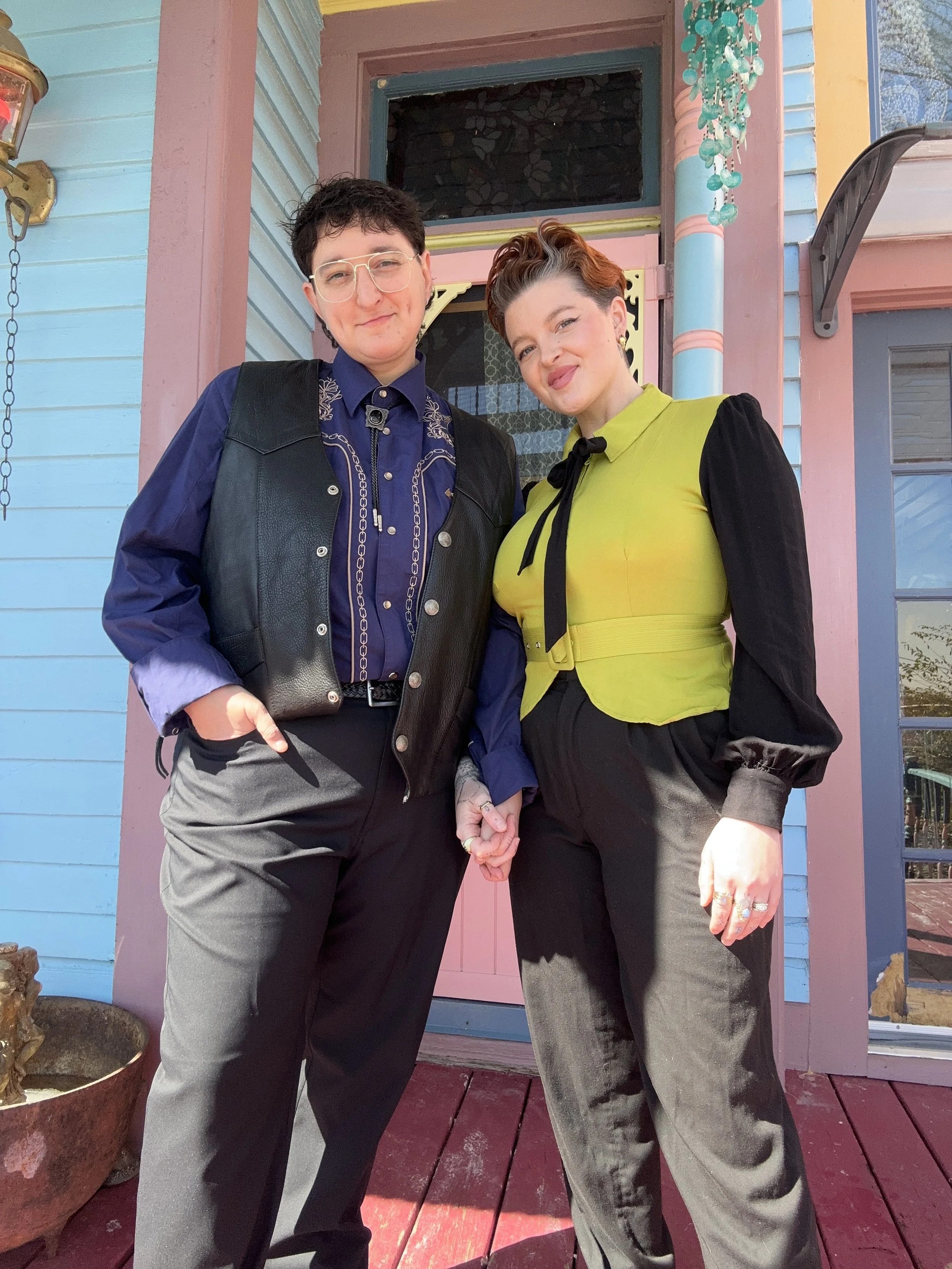 Two people standing close together outside a colorful house, holding hands. One person is wearing a dark vest and glasses, while the other wears a bright green top with black accents. The house behind them is painted in pastel shades of blue, pink, and yellow, with decorative trim and a hanging ornament of green beads.