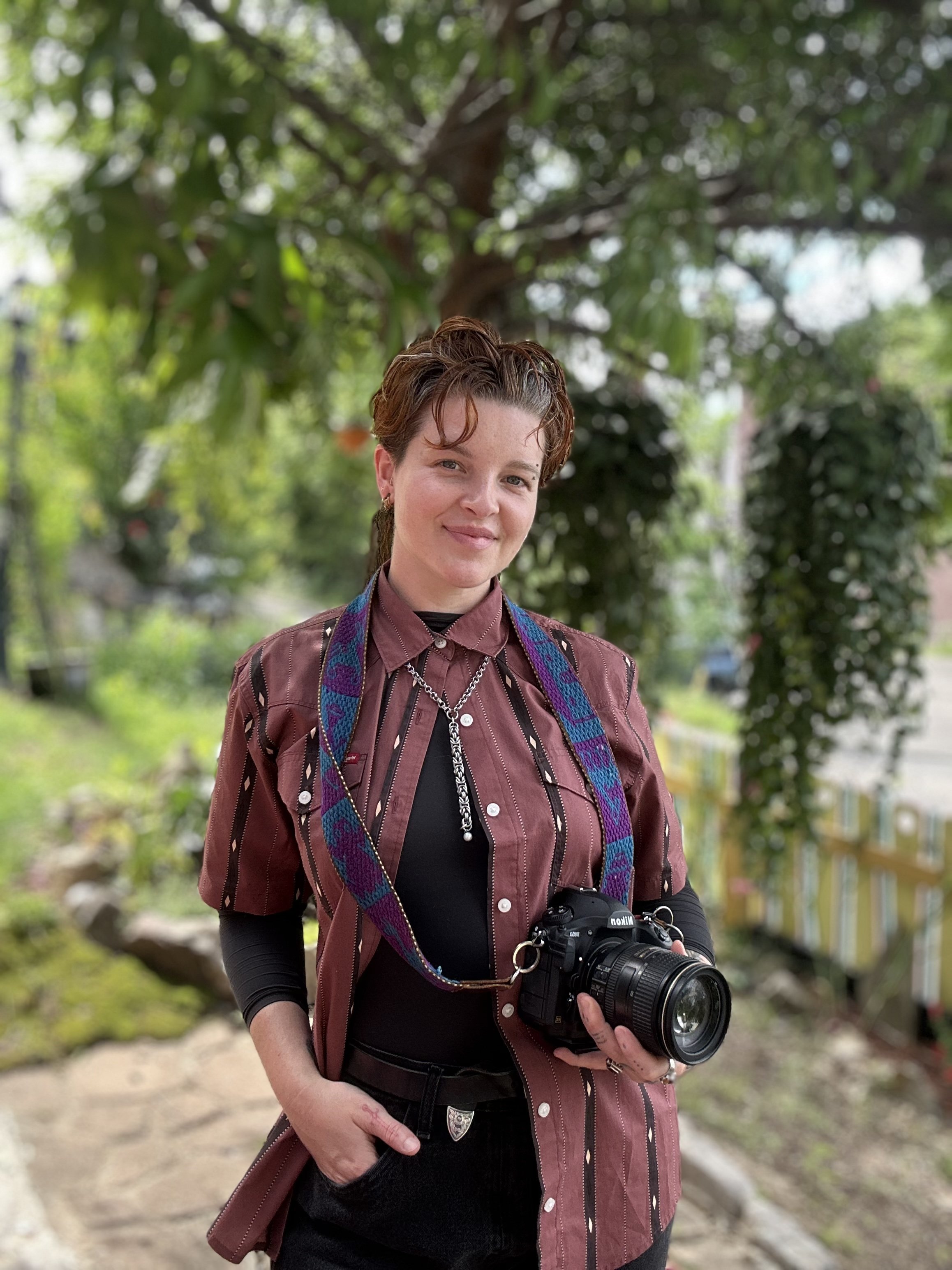 A young woman with short, curly hair and earrings, holding a professional camera, standing outdoors in front of a large tree with green foliage.