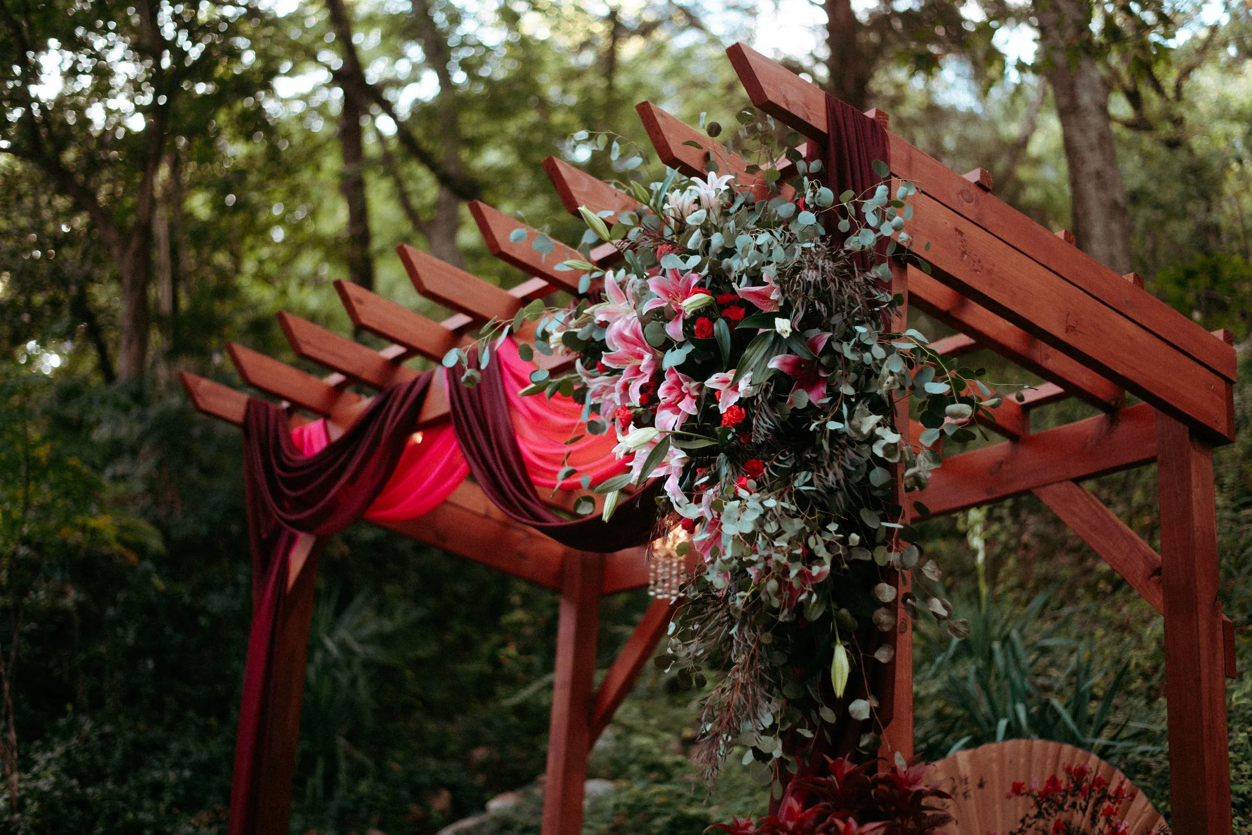 Flower arrangement with pink lilies, red roses, and greenery on a wooden arch at an outdoor setting.